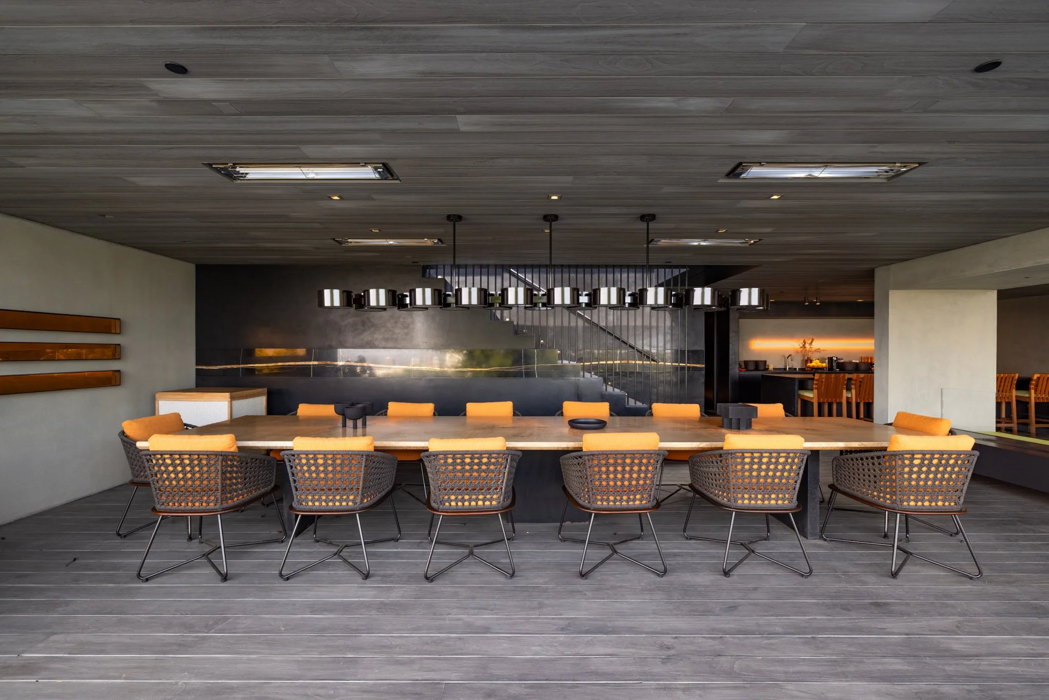 Modern dining room with a long wooden table, black wicker chairs with orange cushions, ceiling lights, and a bar area in the background.