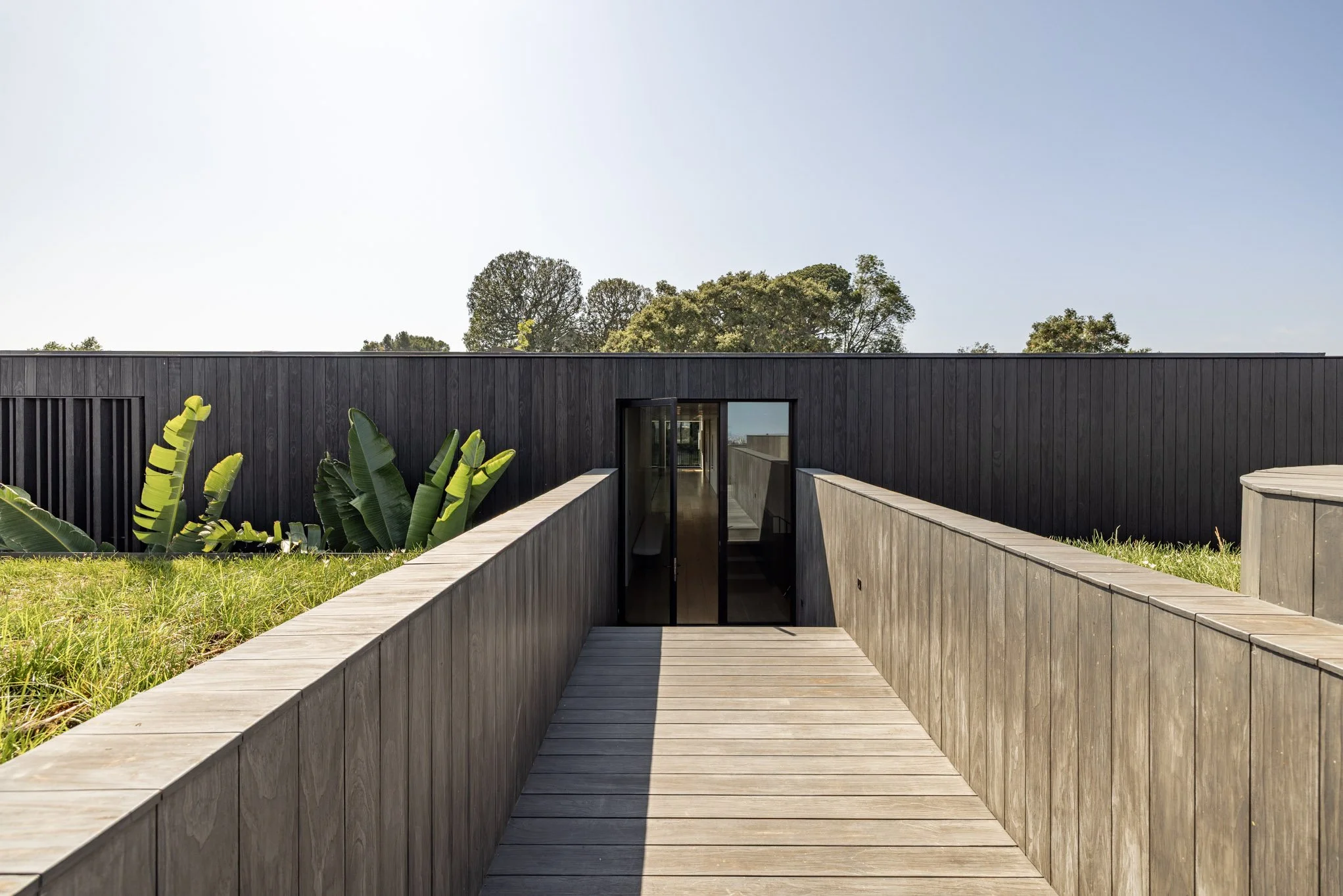Wooden walkway leading to a modern black building with large glass door, surrounded by grassy area and tropical plants, with a bright blue sky and trees in the background.