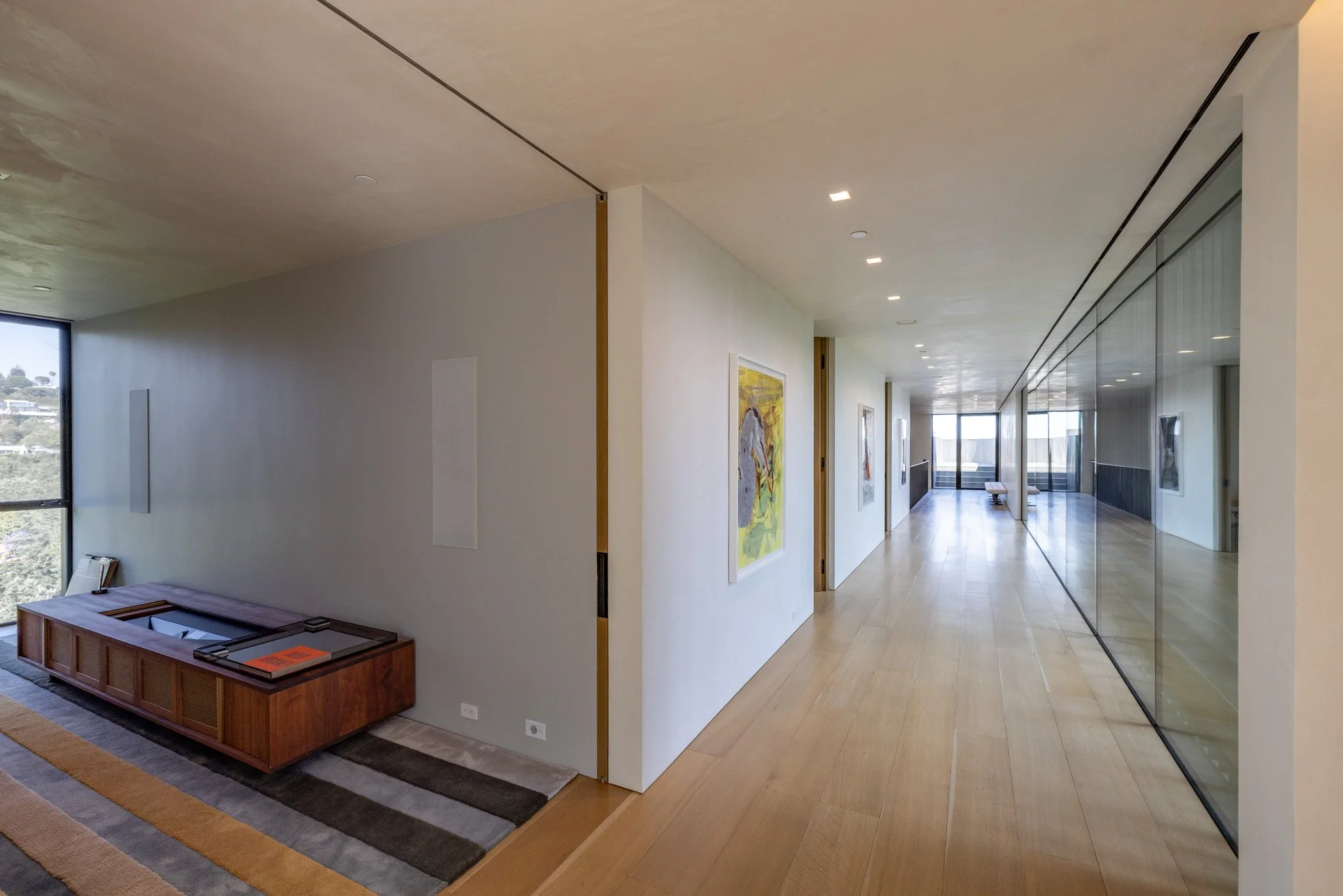 Modern hallway with wooden flooring, white walls decorated with colorful artwork, and a large glass partition on the right reflecting the hallway. At the end, there is a sitting area with large windows letting in natural light.
