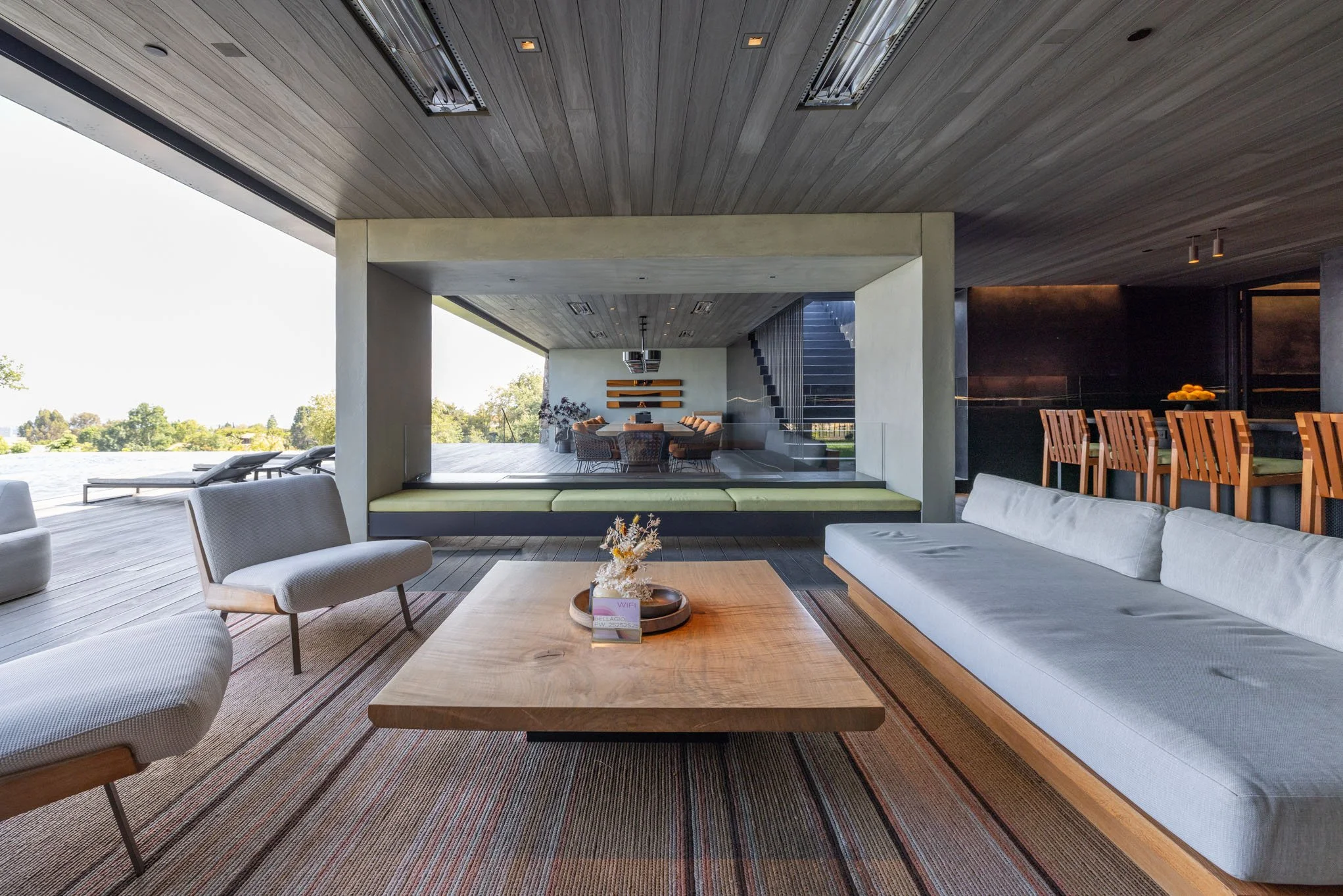 Modern living room with large sofa, armchairs, wooden coffee table, and indoor-outdoor flow overlooking a patio and pool, with stairs and seating area inside.