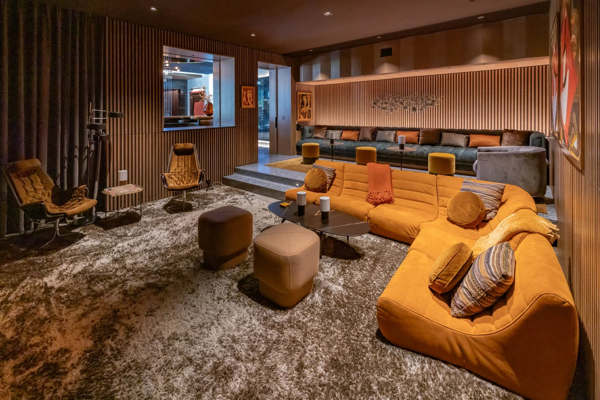 A stylish living room with a large yellow sectional sofa, a black coffee table, and various armchairs, set against wooden paneled walls with modern artwork and artwork on the walls, with dim lighting and plush carpet flooring.