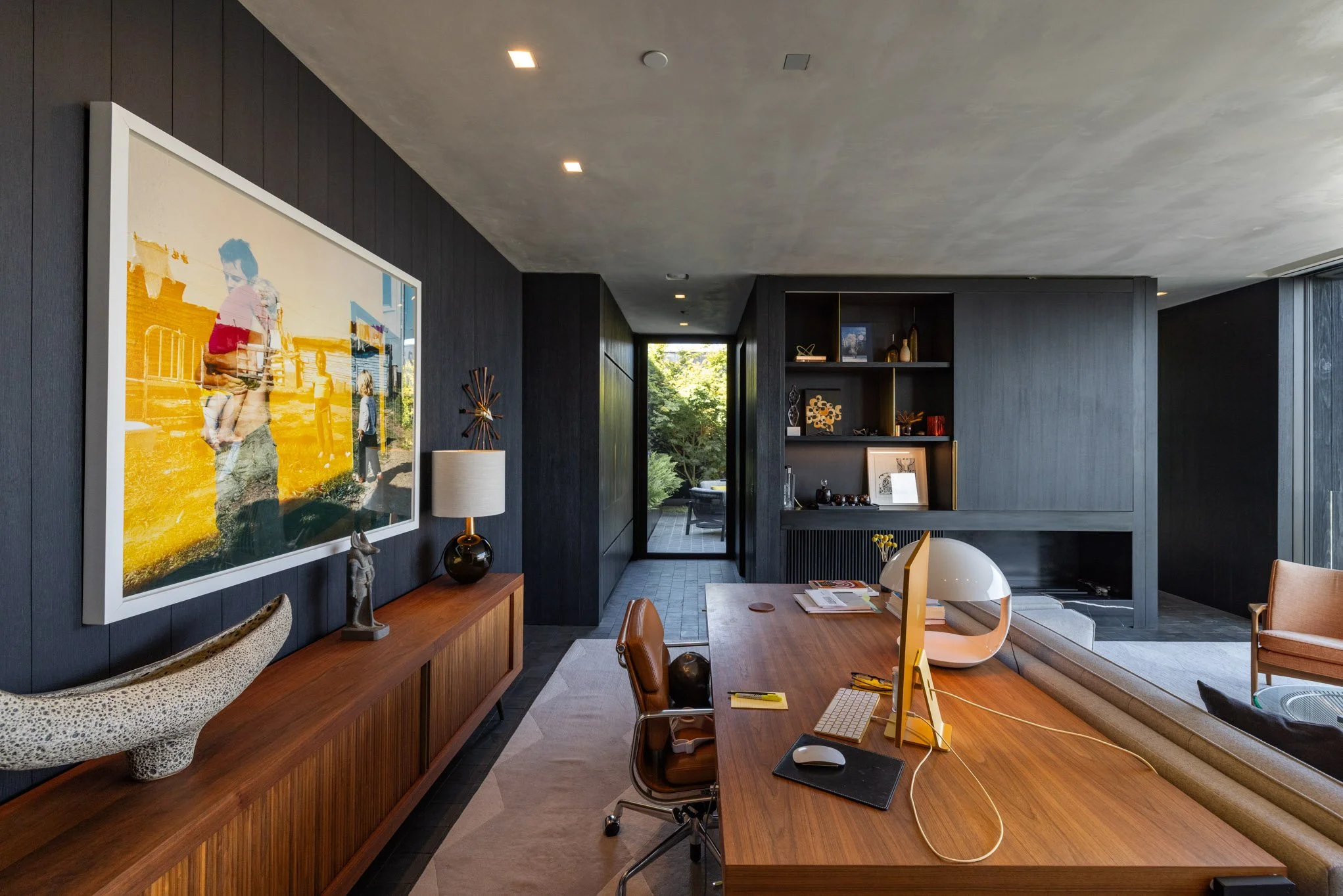 Modern home office with dark wood walls and shelves, a large wooden desk with a computer and mouse, a tan leather office chair, a large abstract vase, a lamp, and a framed picture, with a view of a patio and greenery in the background.
