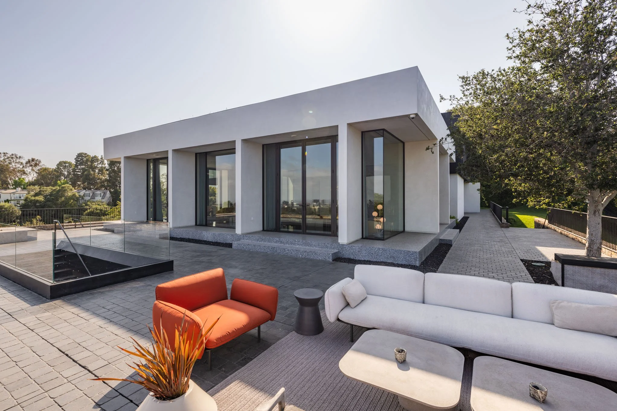 Modern white house with large glass sliding doors, outdoor patio furniture including an orange couch, white sofa, and small black side table, surrounded by trees and a paved walkway.