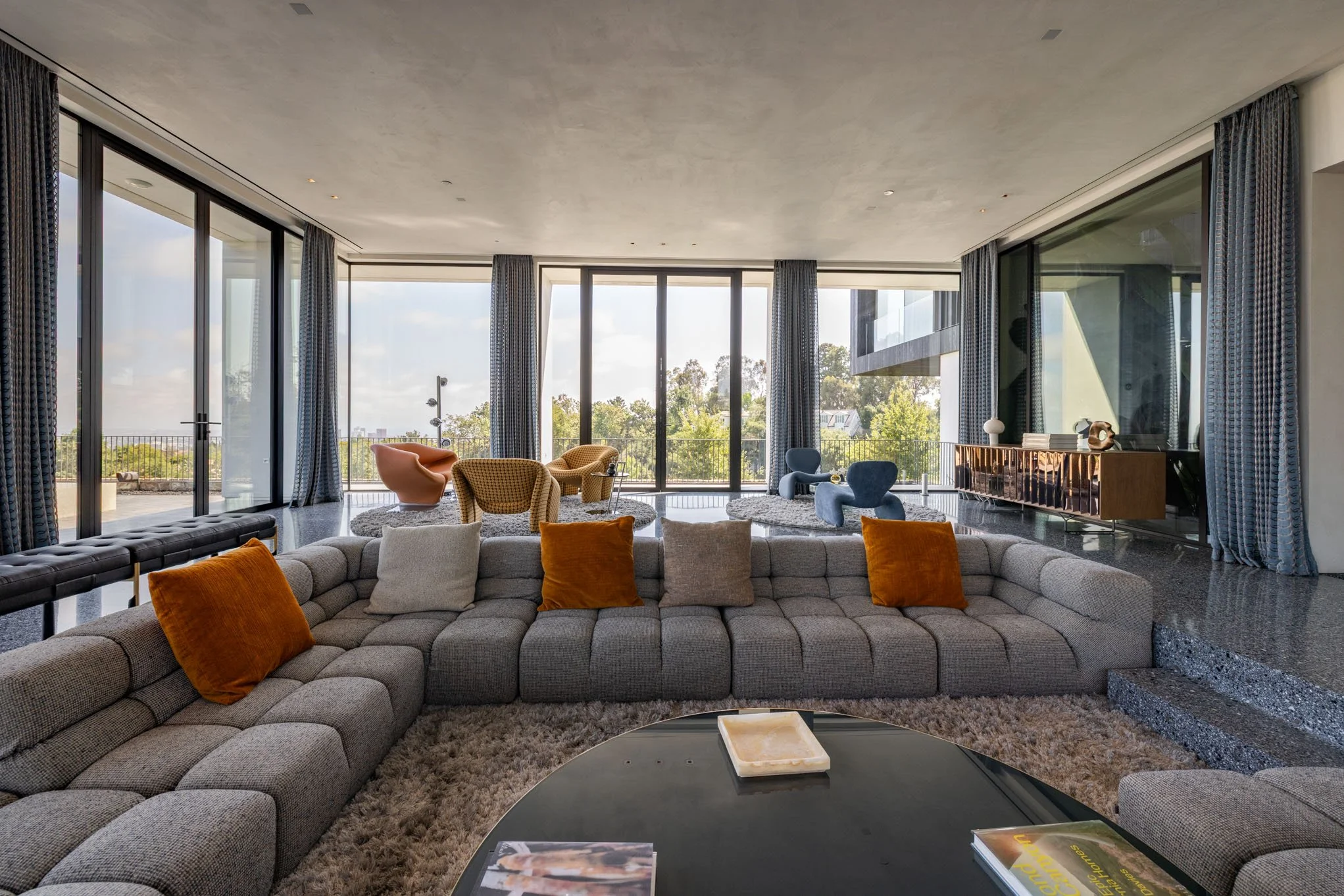Modern living room with a large gray sectional sofa with orange and beige pillows, a round black coffee table with magazines and a decorative box, and several colorful armchairs near floor-to-ceiling windows that let in natural light and show a view 