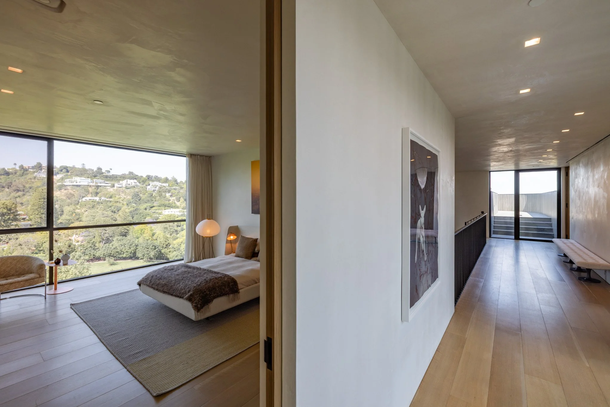 Interior view of a modern apartment or hotel room with a large window, a bed with pillows and a blanket, bedside lamps, a small table with decorative items, and a sitting area. The hallway has wooden flooring, a framed artwork on the wall, and a benc