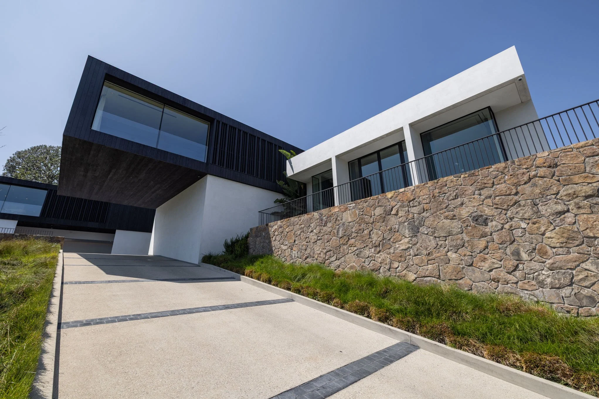 Modern house with black and white exterior walls, large glass windows, stone retaining wall, and a sloped concrete driveway.