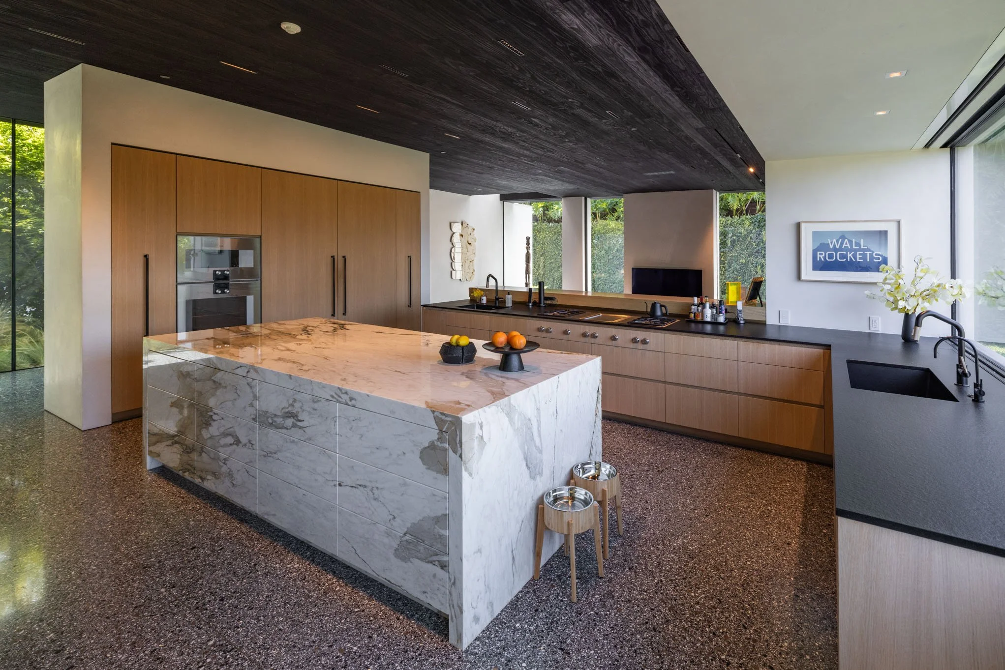 Modern kitchen with a marble island, wooden cabinetry, dark countertop, black sink, and large windows showing greenery outside.
