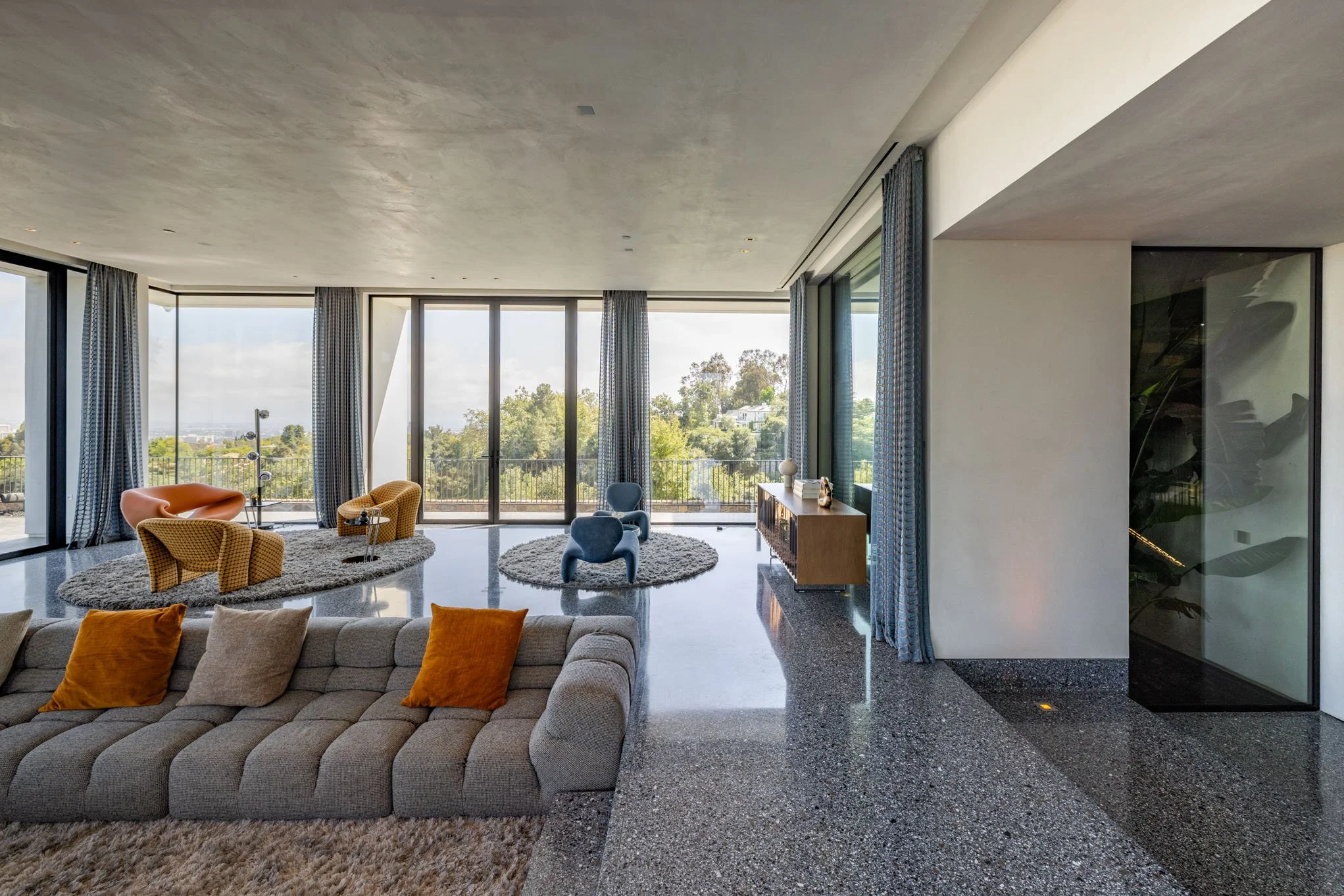 Modern living room with large windows, beige sofa with orange and beige pillows, yellow and blue accent chairs, glass sliding doors, and a view of greenery outside.