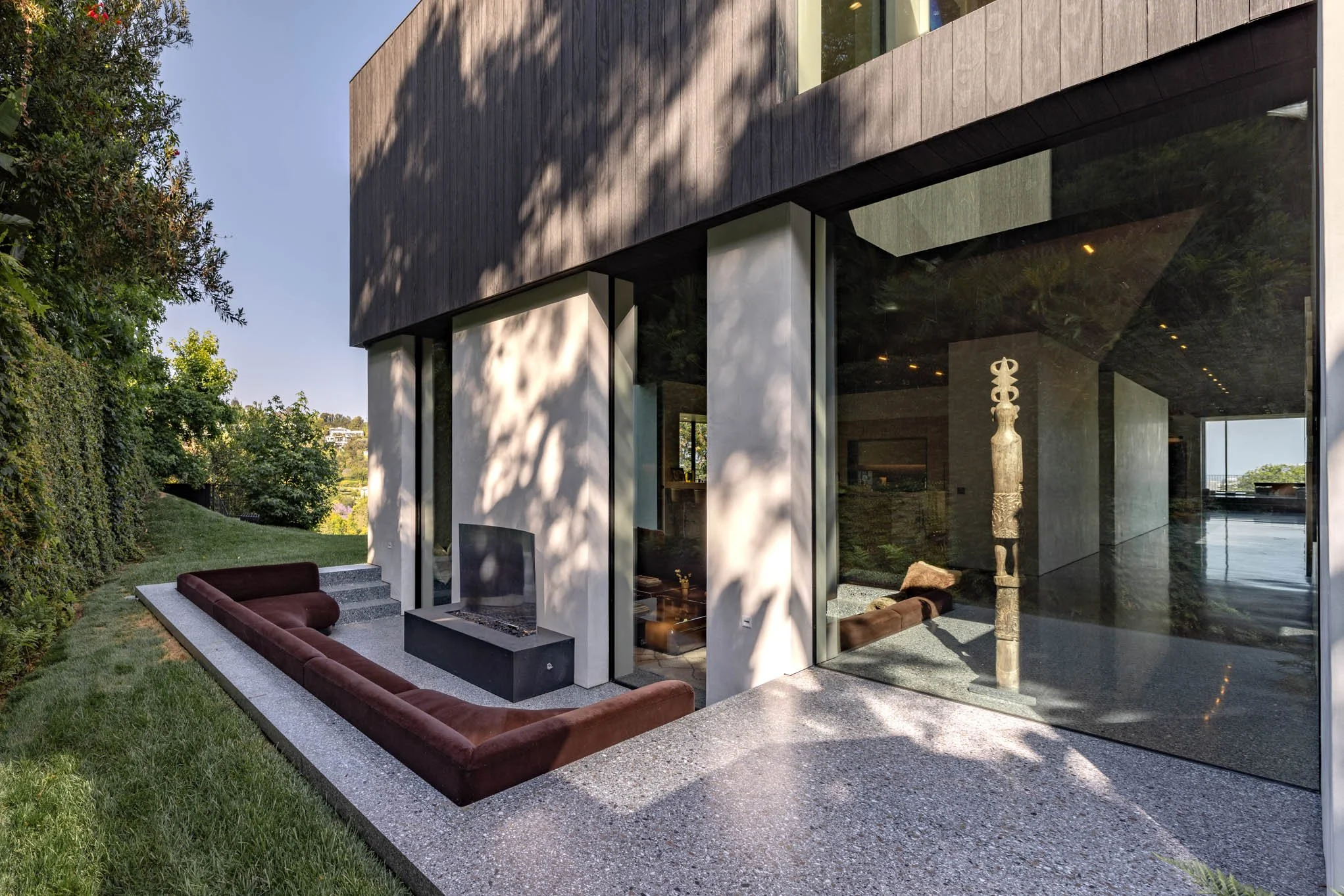 Modern house with large glass windows, an outdoor fire pit, long upholstered seating, and minimalist decor surrounded by greenery.