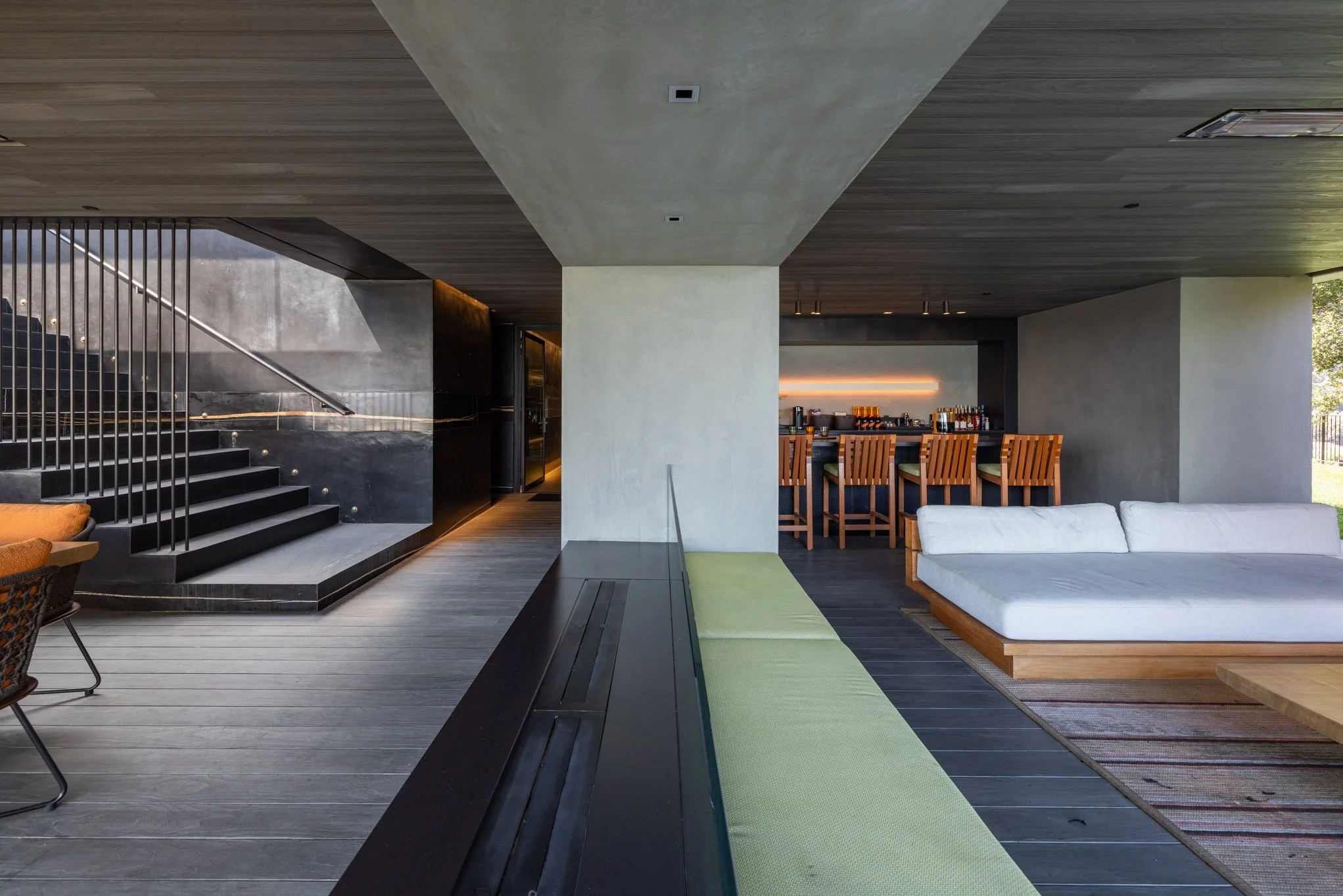 Modern living room with a staircase, bar area, and seating area featuring a white sofa, green bench, and bar stools, with wooden and black accents.