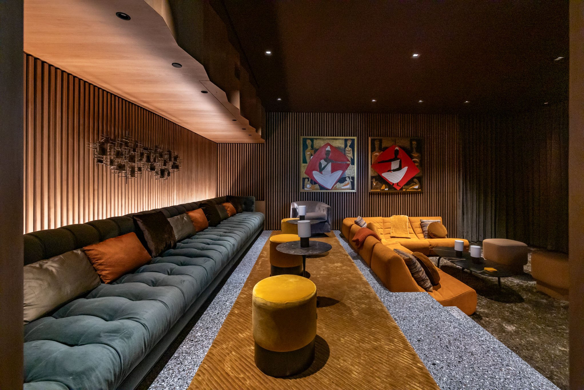 Modern lounge area with long black couch, yellow ottomans, and a mustard-colored sofa, featuring artistic paintings on wooden-paneled walls and dim ambient lighting.