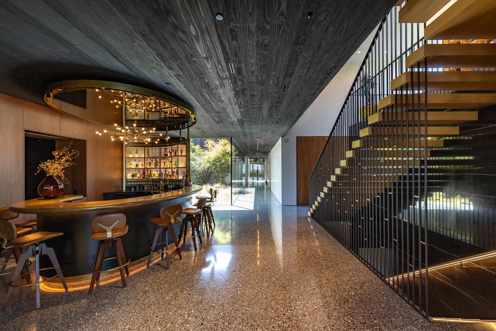 Modern bar with curved wooden counter, barstools, and a black metal staircase in a stylish, well-lit interior with a large window. Decor includes a vase with branches and artistic lighting fixture.