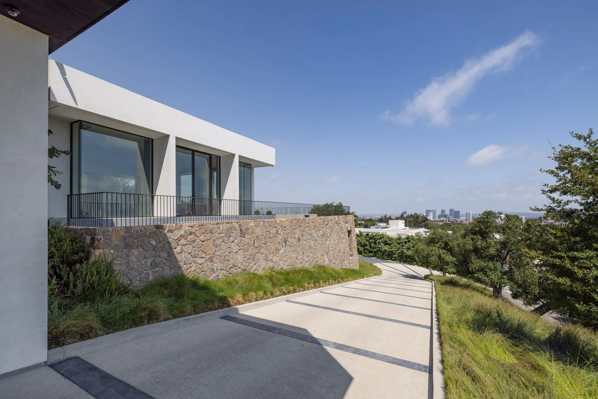 Modern house with large glass windows, stone wall, and patio overlooking a city skyline with trees and a partly cloudy sky.