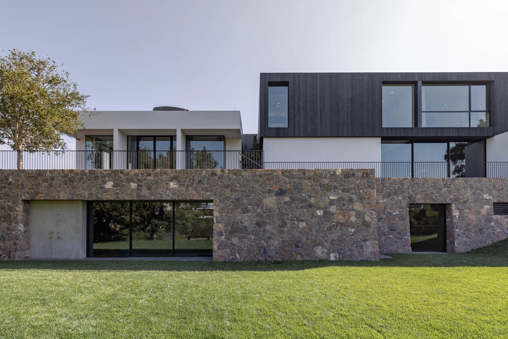 Modern multi-story house with a stone and white facade, large glass windows, and a green lawn in the foreground.