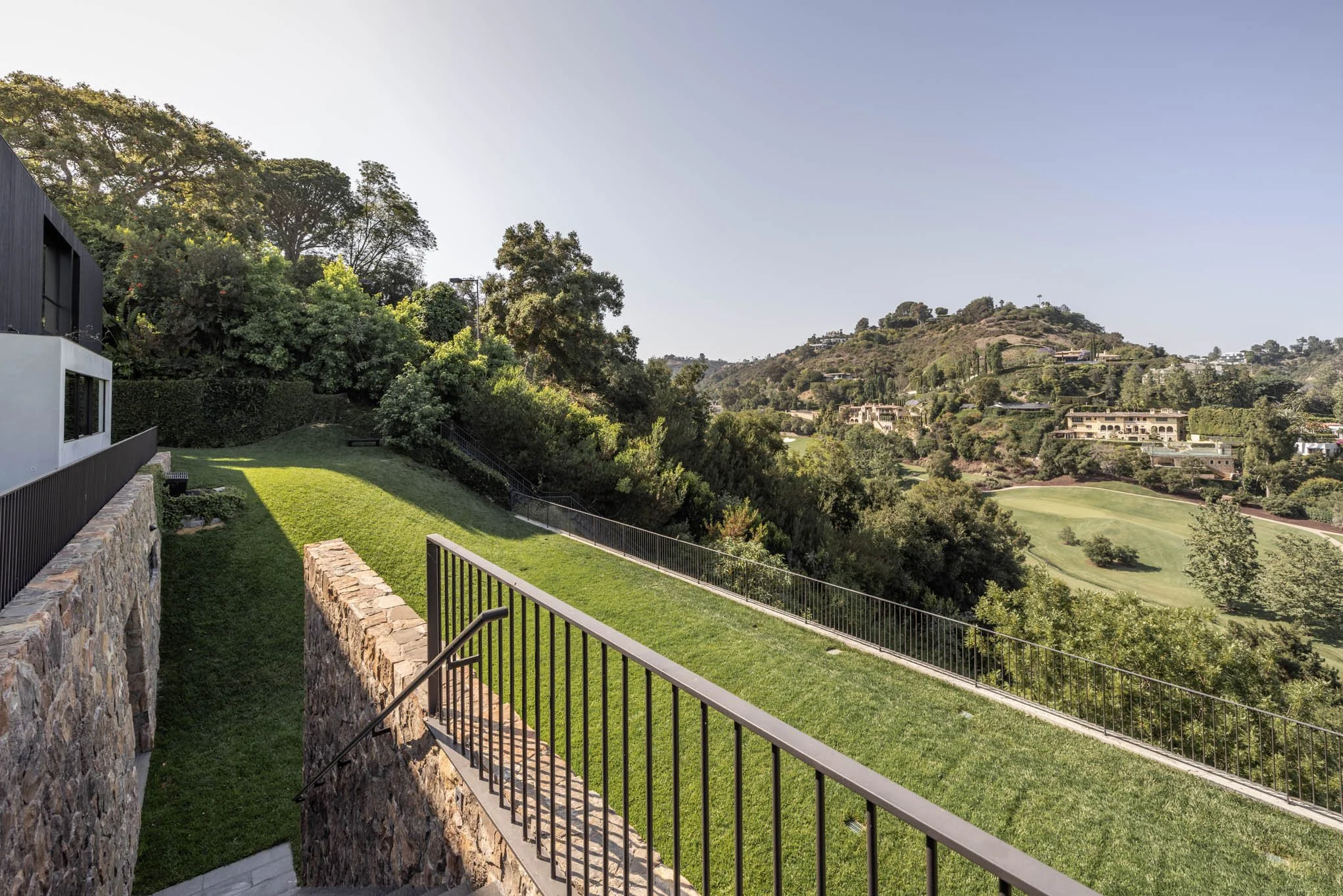 Scenic view of a hillside with houses, trees, and a golf course in the distance seen from a patio with a stone and metal railing.