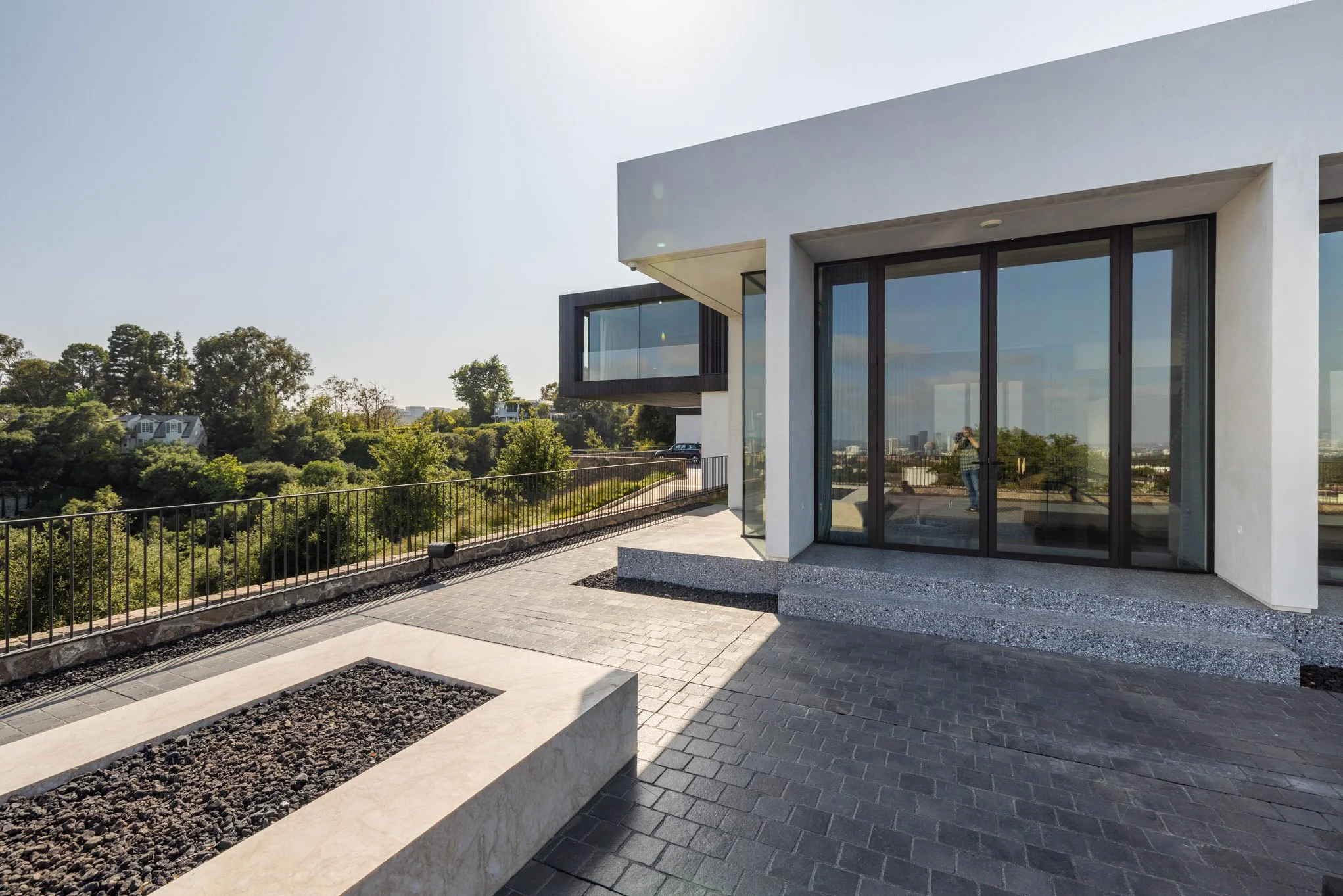 Modern house with large glass doors and a terrace, overlooking trees and a distant city skyline on a sunny day.