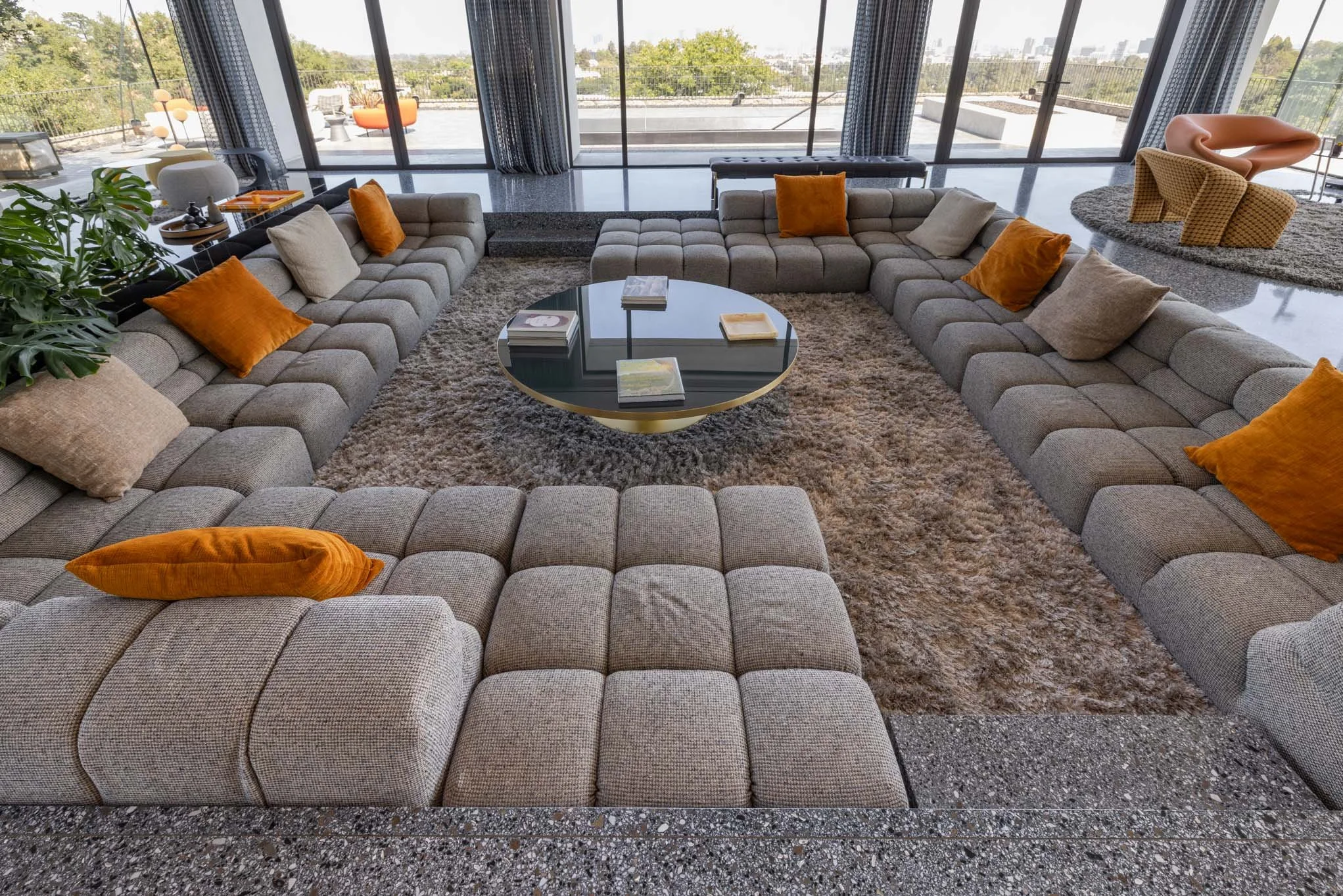 Contemporary living room with a large gray sectional sofa, orange and beige pillows, a round glass coffee table, a shaggy beige rug, and floor-to-ceiling windows showing a balcony with city views.