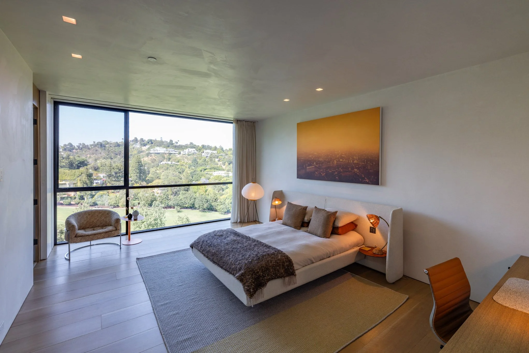 Modern bedroom with a large floor-to-ceiling window overlooking a green hillside, a white bed with beige and brown pillows, a fur blanket, two bedside lamps, a wall art depicting a cityscape at sunset, a chair by the window, and a wooden desk with a 