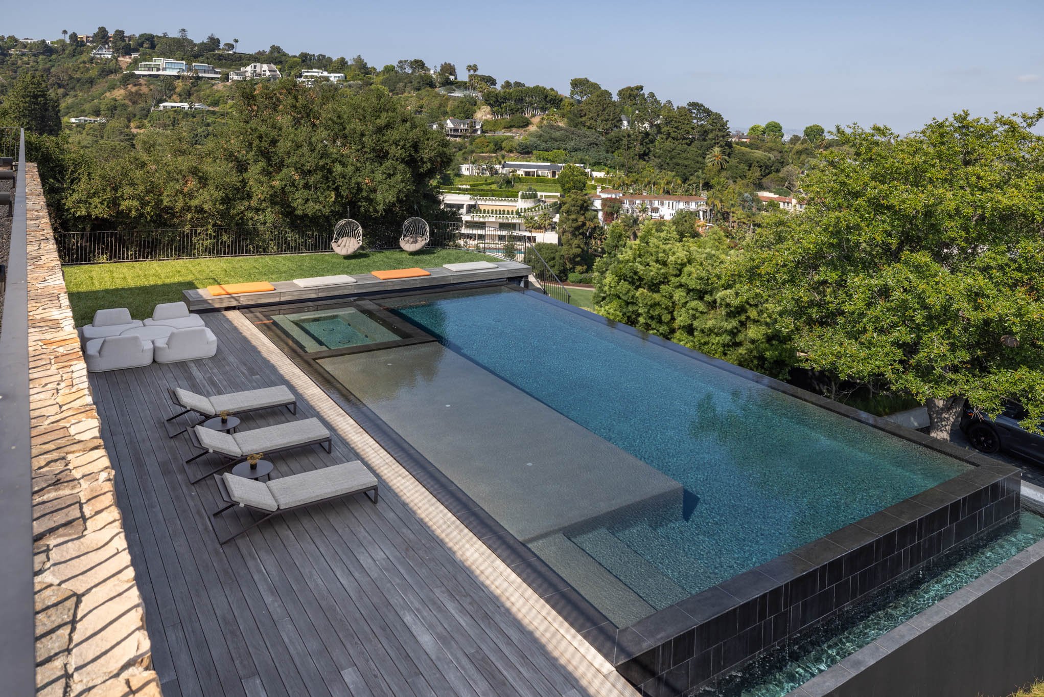 A modern rooftop infinity pool with a wooden deck, lounge chairs, and outdoor seating, overlooking a lush green neighborhood with trees and houses on a hillside.