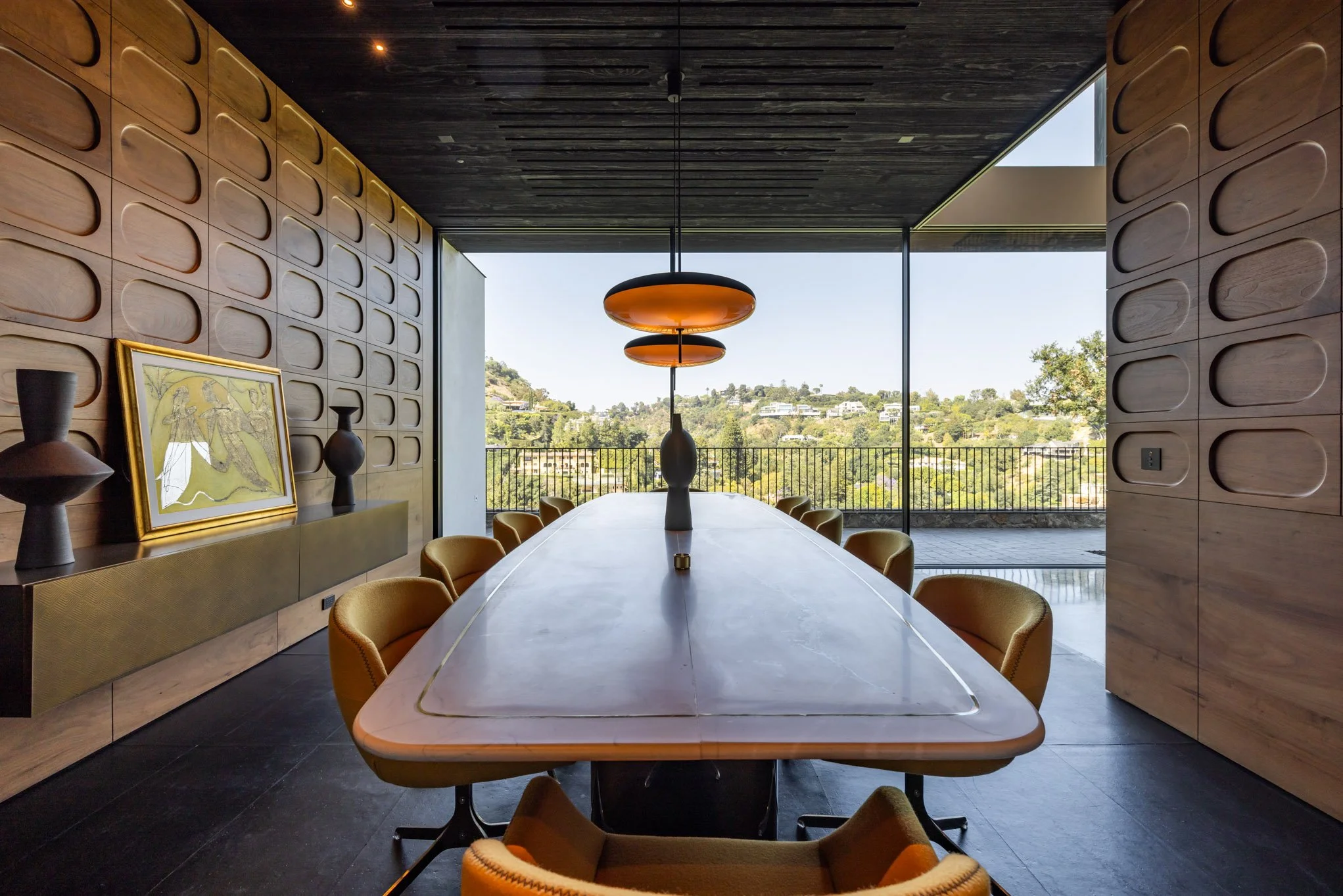 Modern dining room with a long oval table, surrounded by mustard-colored chairs, featuring wood paneled walls, a large window with a view of greenery, and stylish orange pendant lights hanging over the table.