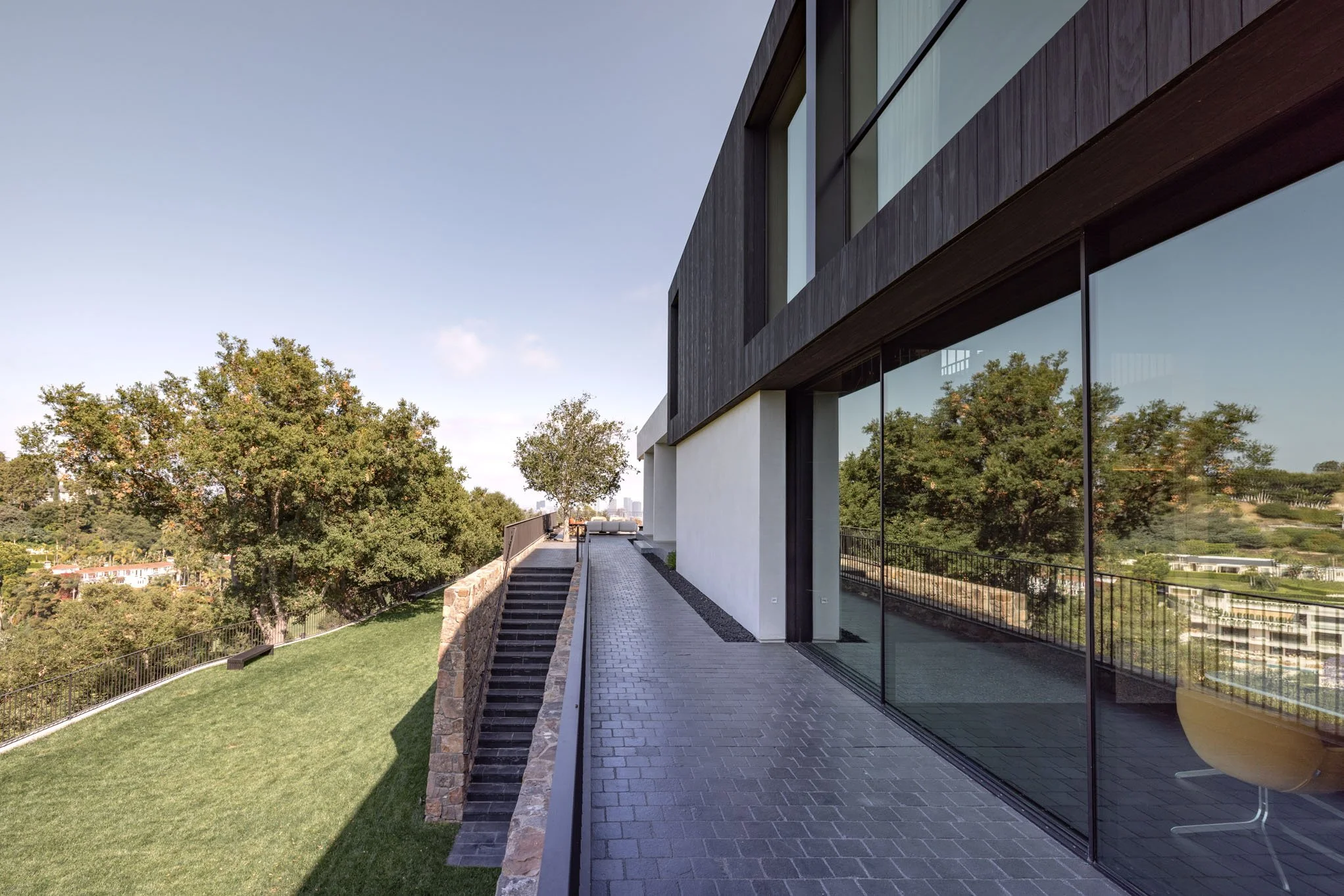 Modern house exterior with large glass windows, black wooden panels, and a paved terrace overlooking trees and a green lawn.