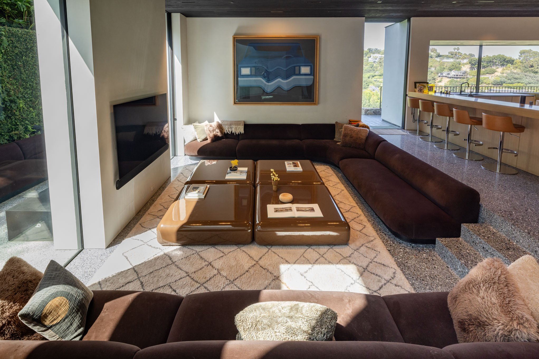 Living room with a large U-shaped dark brown sectional sofa, a modern glossy brown coffee table with books and decor, a wall-mounted TV, a large abstract painting, a patterned area rug, and floor-to-ceiling windows showing an outdoor landscape with g