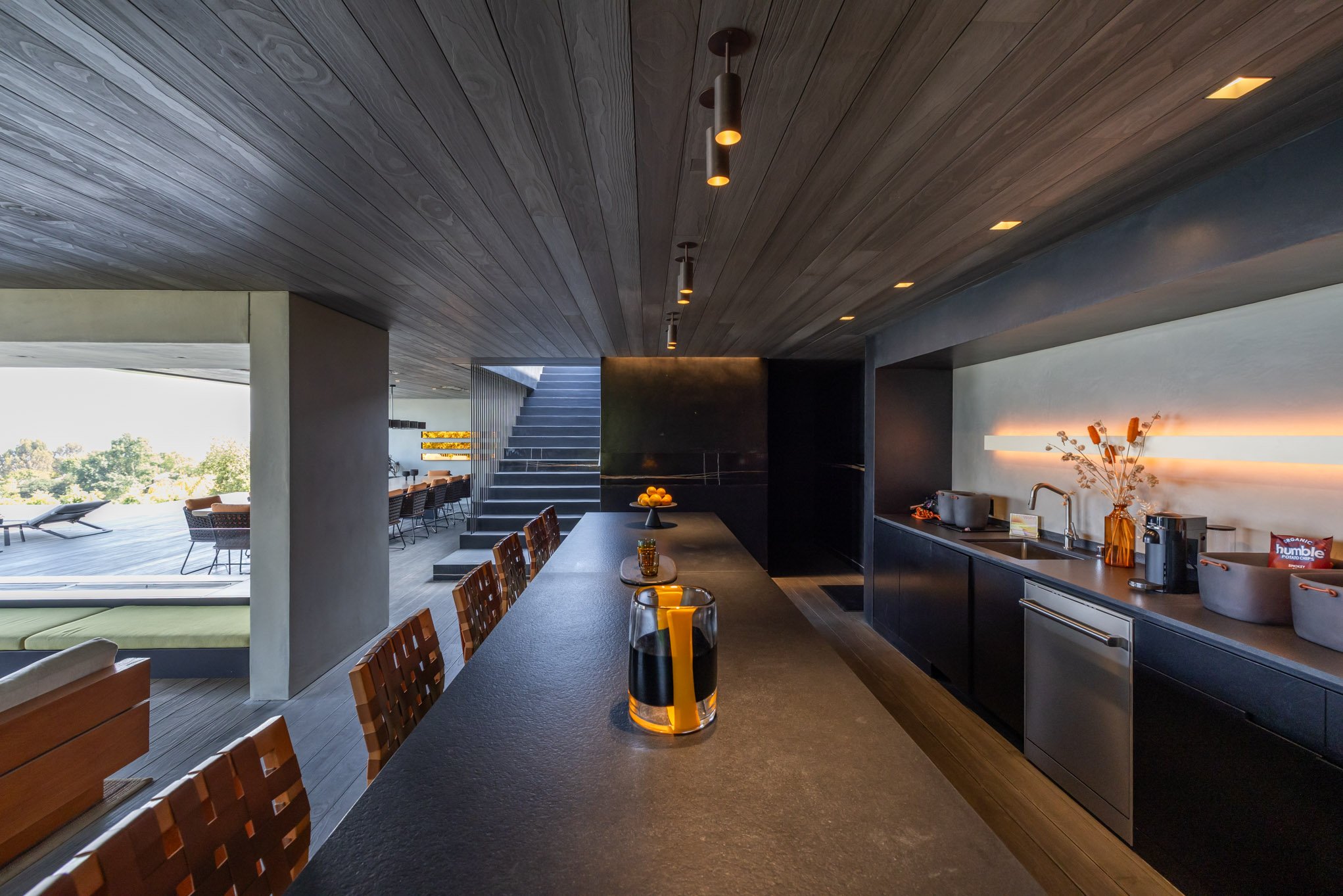 Modern outdoor kitchen and dining area with dark cabinetry, a long black countertop, and wooden chairs, overlooking a patio with lounge chairs and a scenic view of trees.