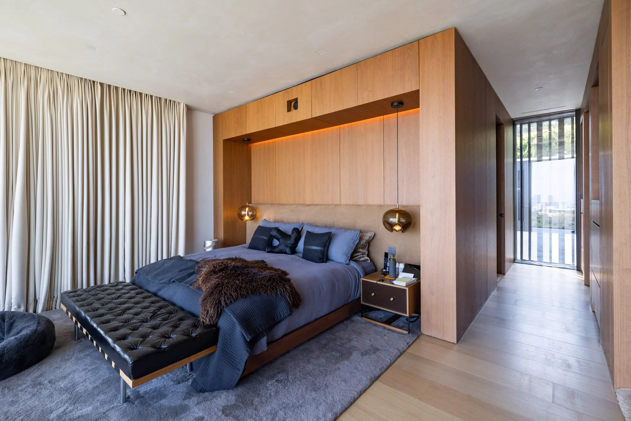 Modern bedroom with a wooden headboard, black and gray bedding, pendant lights, a small nightstand with books, and large cream curtains.