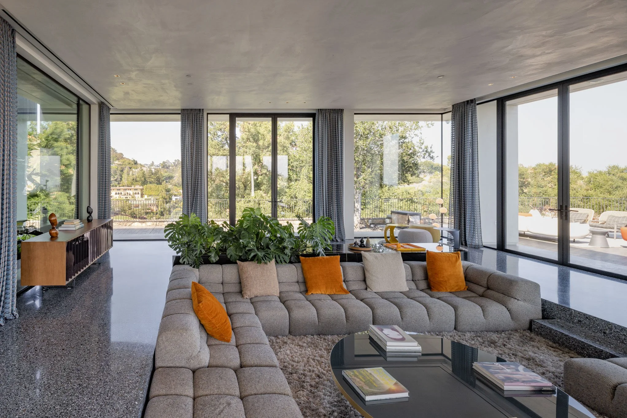 Modern living room with large glass windows, beige sectional sofa with orange and beige throw pillows, a black coffee table with books, and greenery outside.