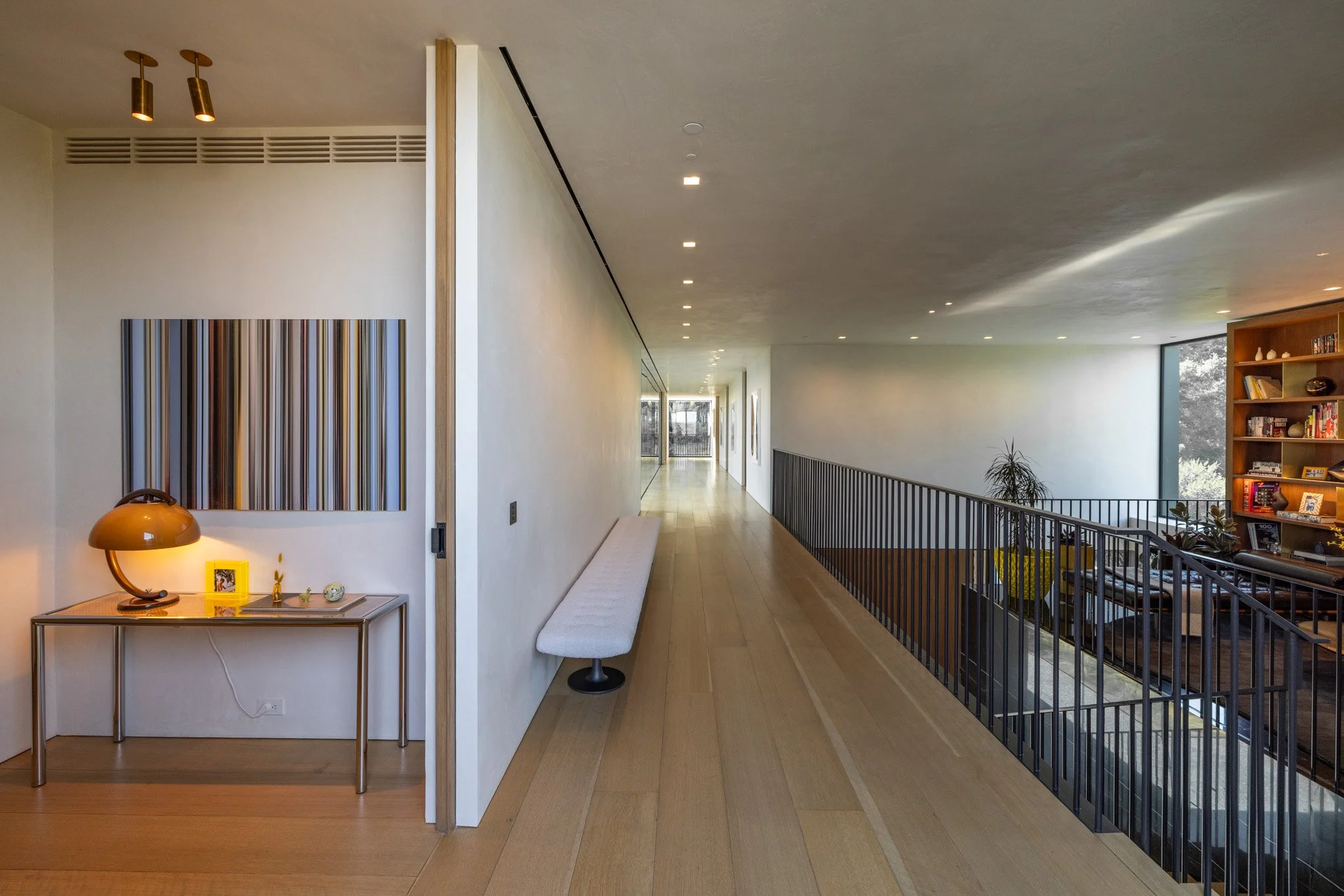 Modern interior hallway with wooden flooring, a long white bench on the left, artwork on the wall, and a staircase with black railing leading to a living space with a bookshelf and large windows showing trees outside.