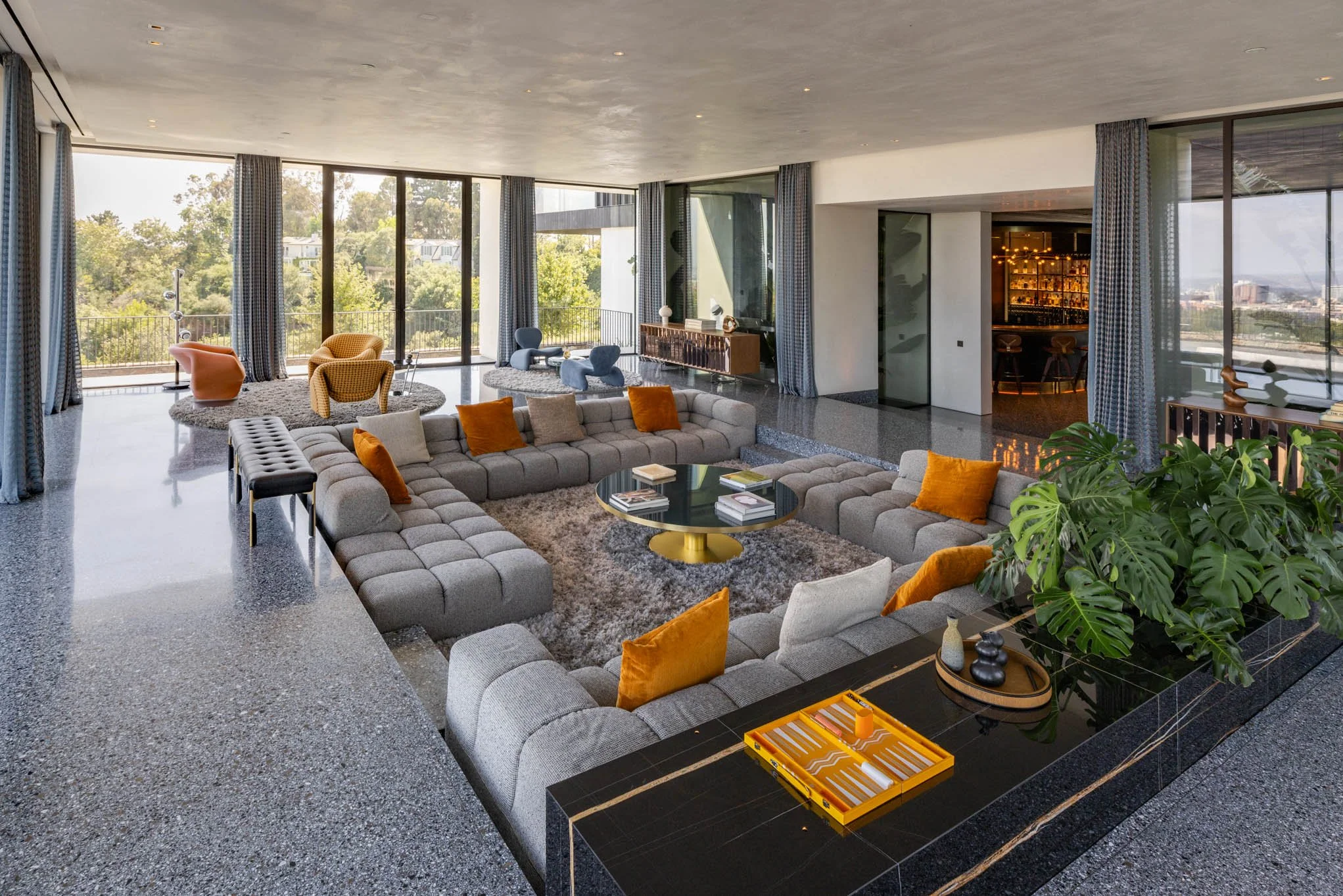 Modern living room with large windows, gray sectional sofa with orange and beige pillows, round coffee table, assorted colorful chairs, indoor plants, and bar area in background