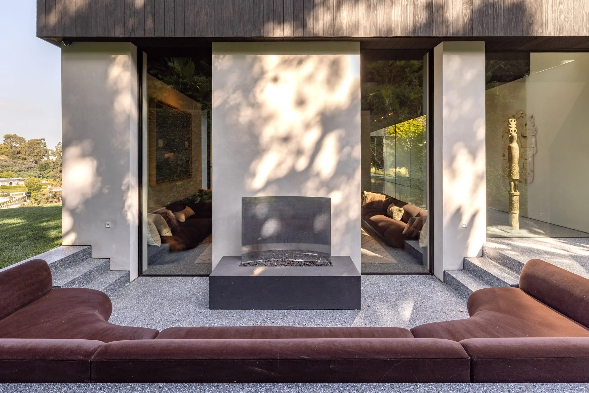Contemporary outdoor seating area with a brown sectional couch arranged in a U-shape, facing a central fireplace, with large glass walls behind and shadows of trees on the walls. Inside, there are more couches and a sculpture inside a modern house.