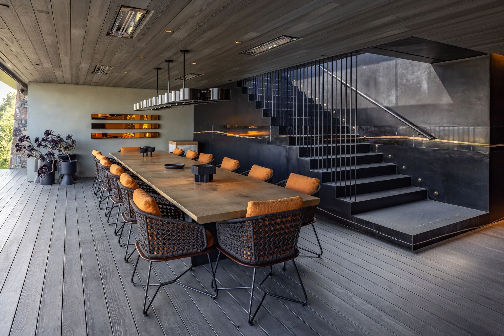 Modern dining room with a long wooden table surrounded by 12 cushioned chairs, black wicker backs, and black metal legs. A staircase with black steps and a thin black railing is located at the back, next to a black wall. On the left, there are potted