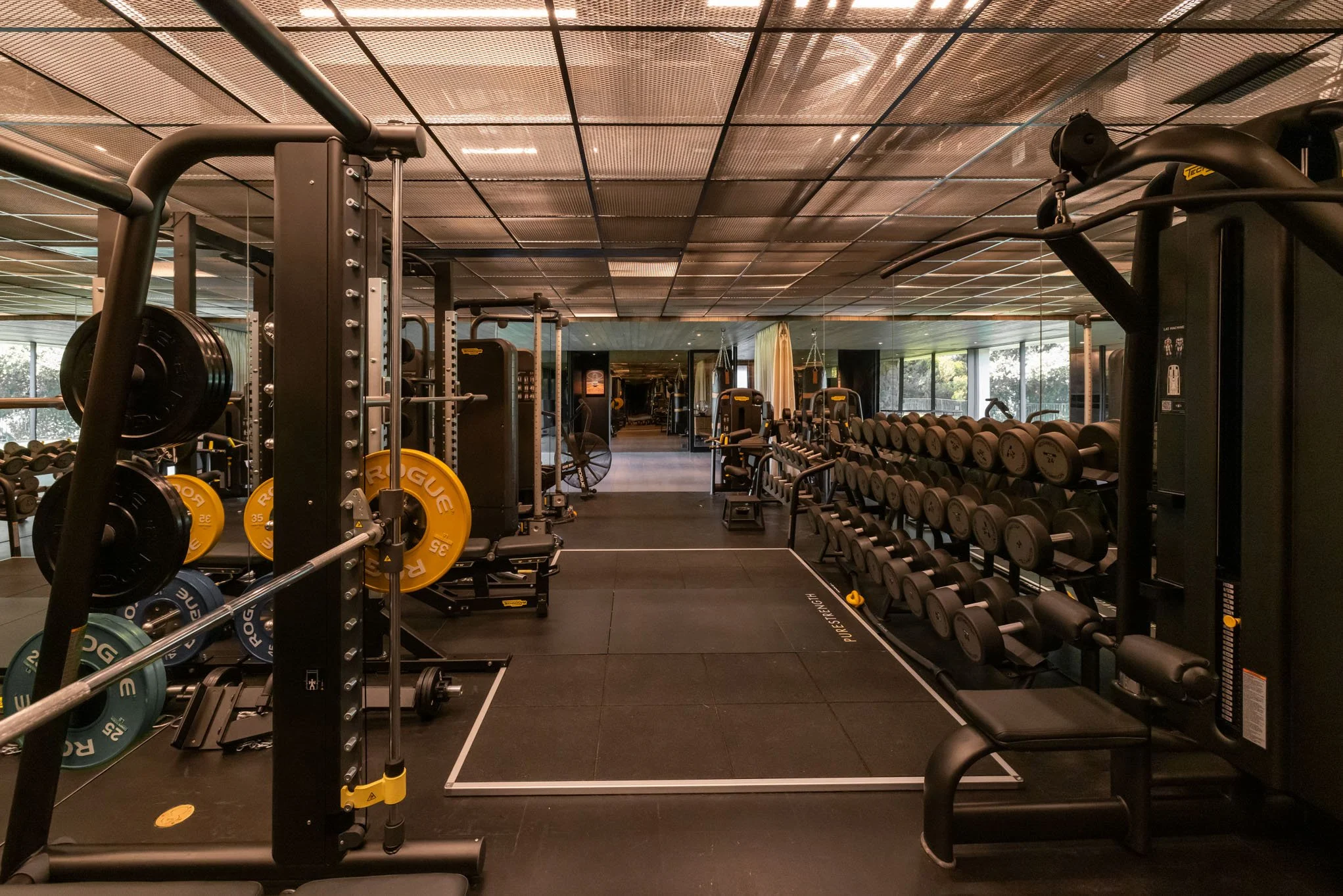 An empty modern gym with weightlifting equipment including dumbbells, barbells, and weight plates, along with cardio machines, in a well-lit space with large windows and a black rubber flooring.