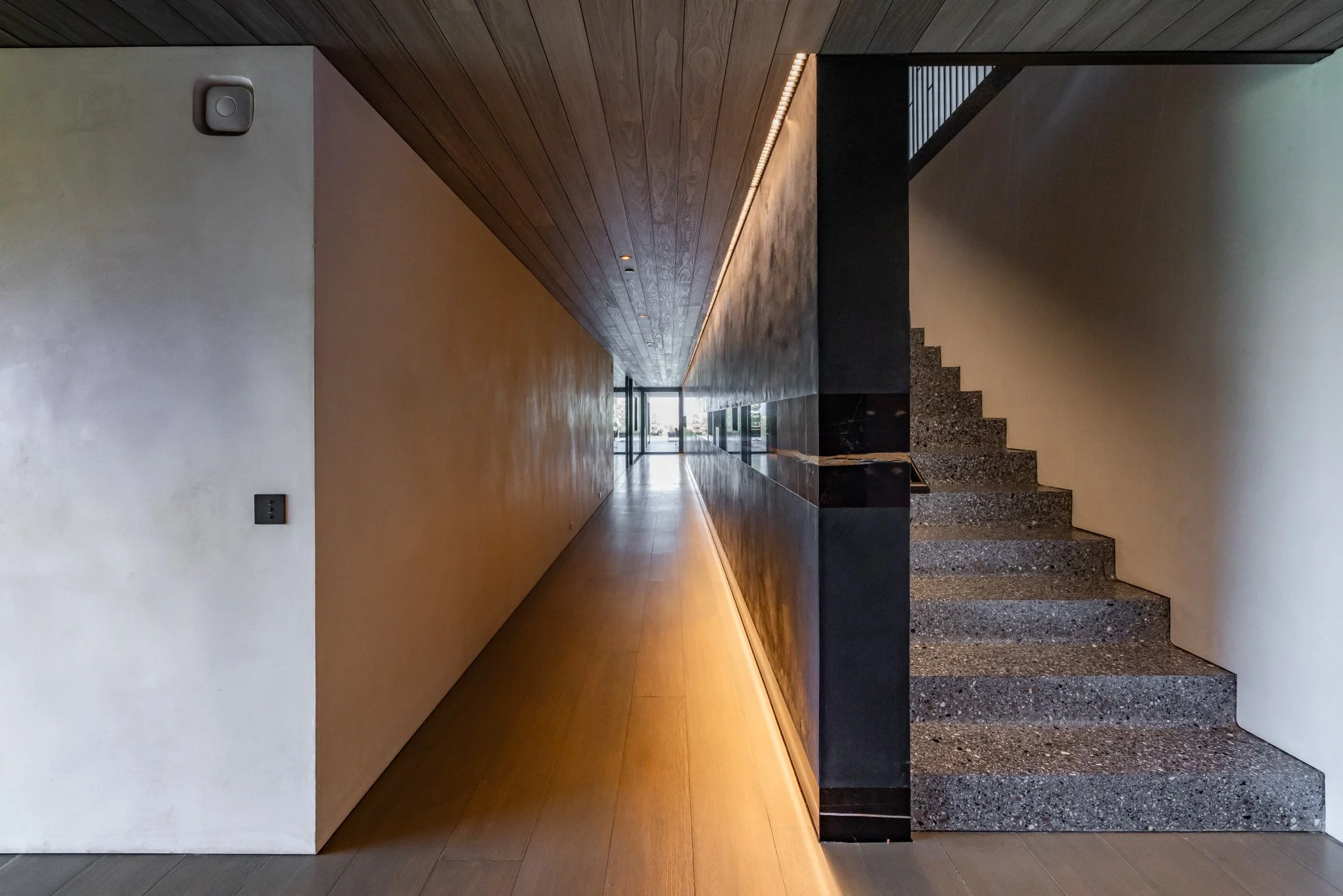 Modern interior hallway with wood ceiling, beige and black walls, staircase, and natural light coming from glass doors at the end.