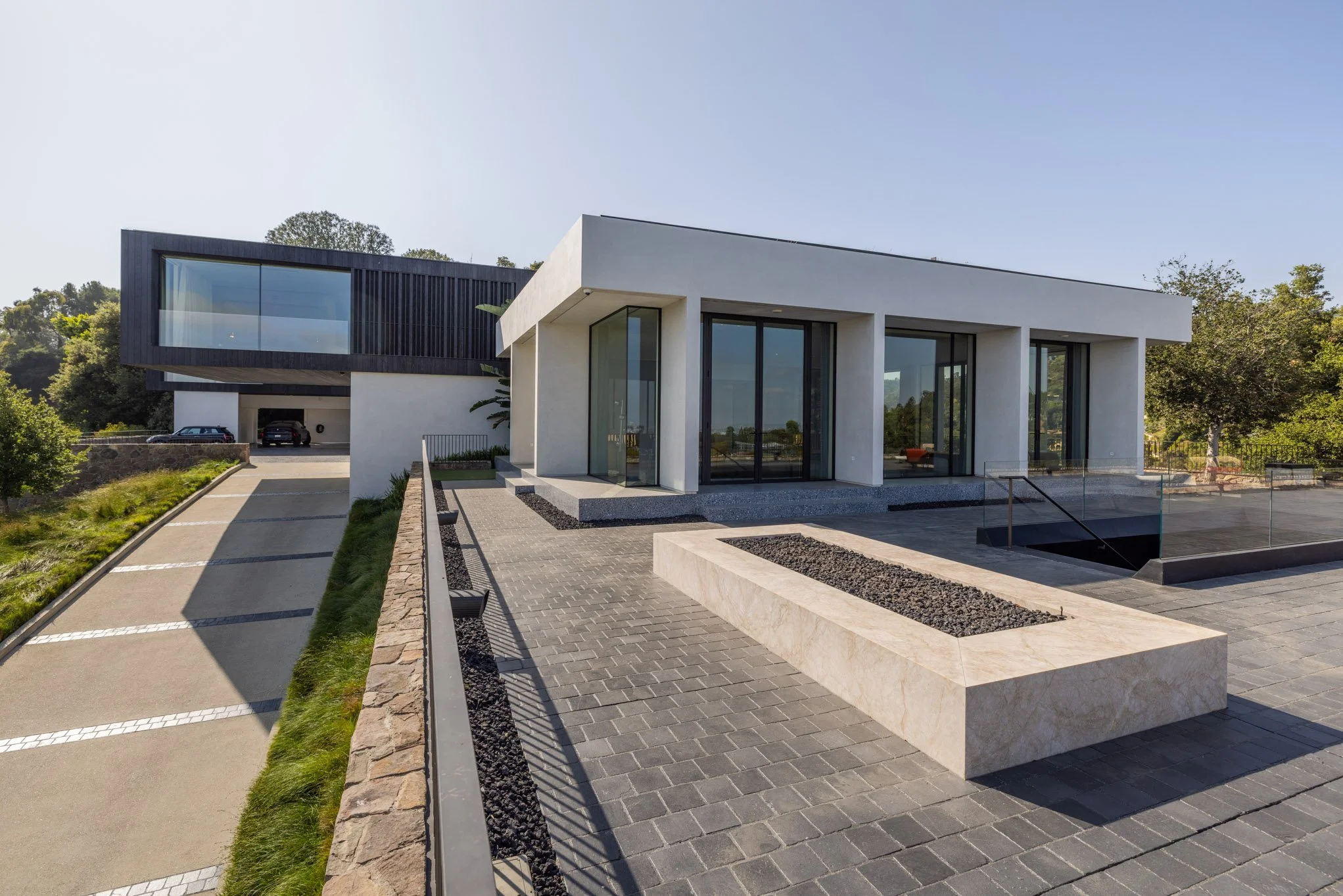 Modern house with white exterior walls, large glass windows, and a black upper section, surrounded by a paved driveway and greenery.