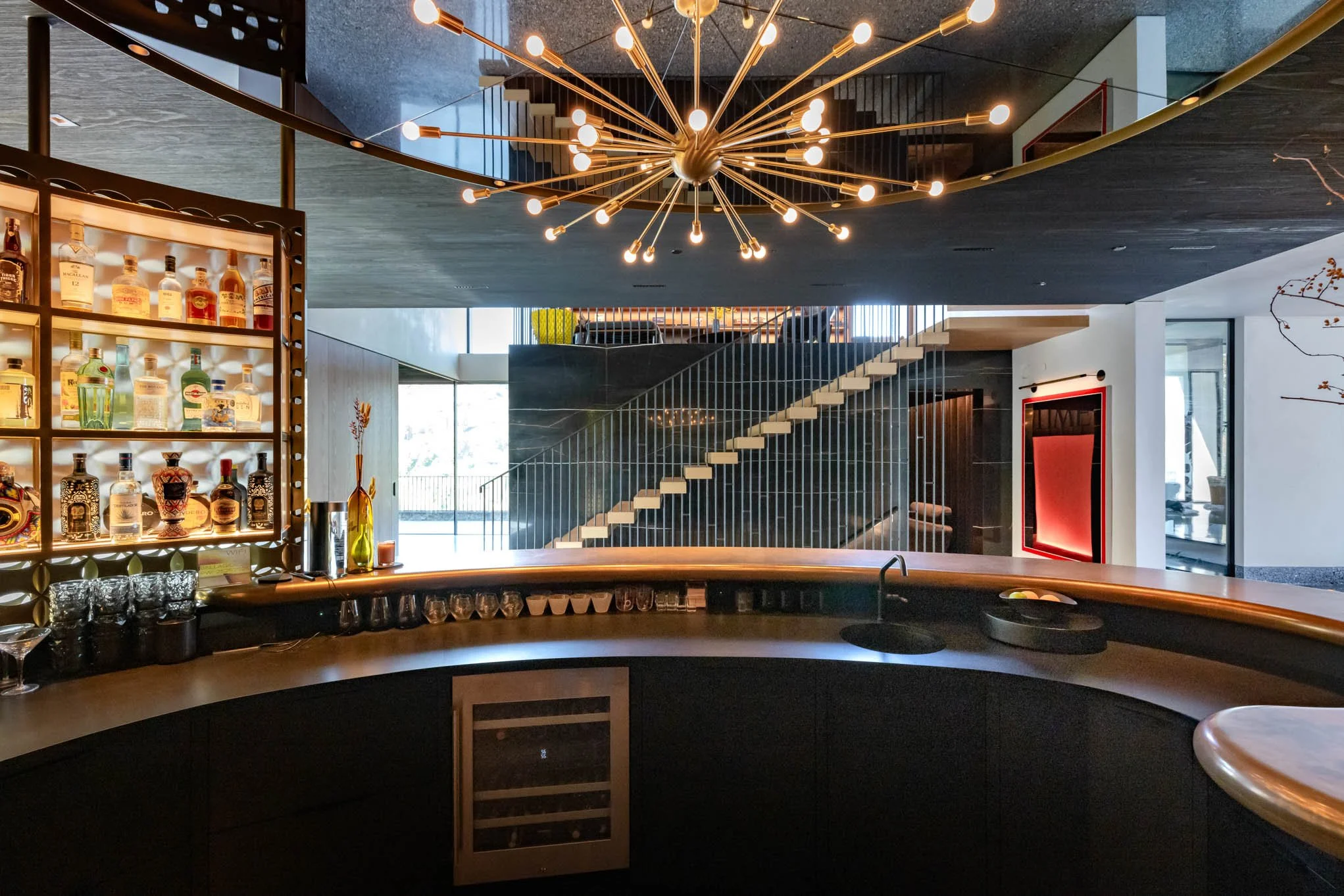 Modern indoor bar area with black curved counter, display of liquor bottles, glassware, and a decorative chandelier with multiple light bulbs, overlooking a staircase with open risers and sleek metal railing.