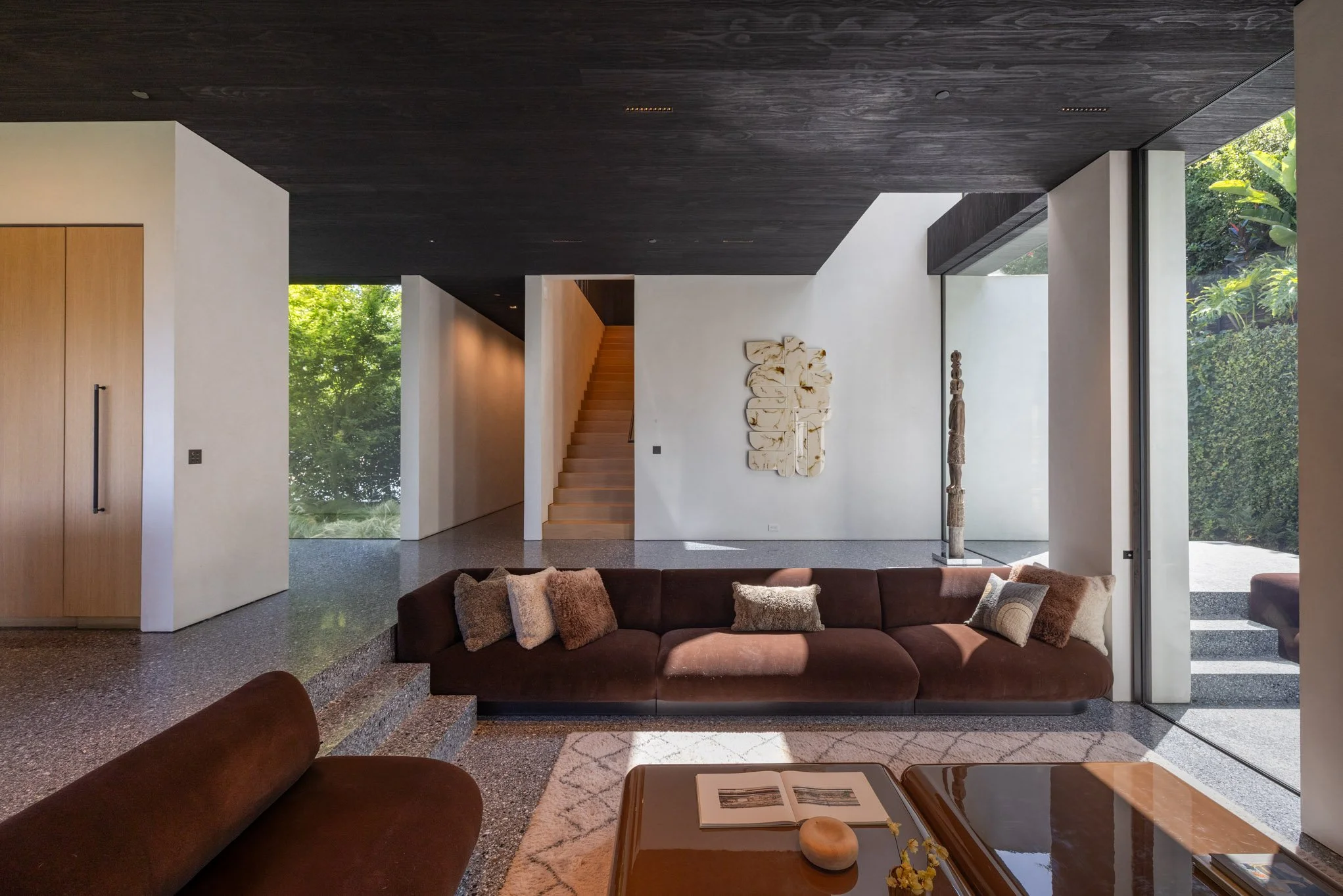 Modern living room with a brown sofa, throw pillows, a wooden coffee table with open book and decorative item, large floor-to-ceiling windows, white walls, dark wood ceiling, staircase, and contemporary art.