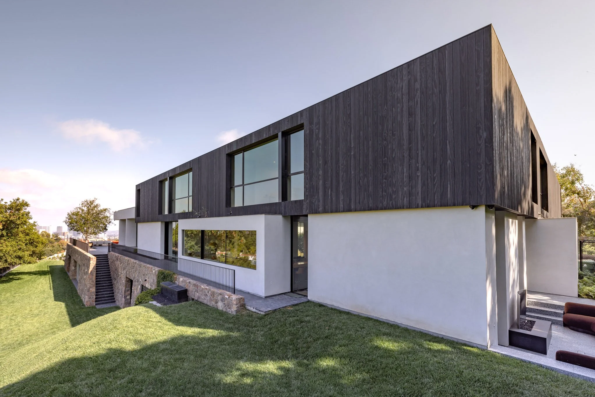 Modern house with a black wooden upper level and white lower level, situated on a grassy slope with a stone staircase and trees in the background.