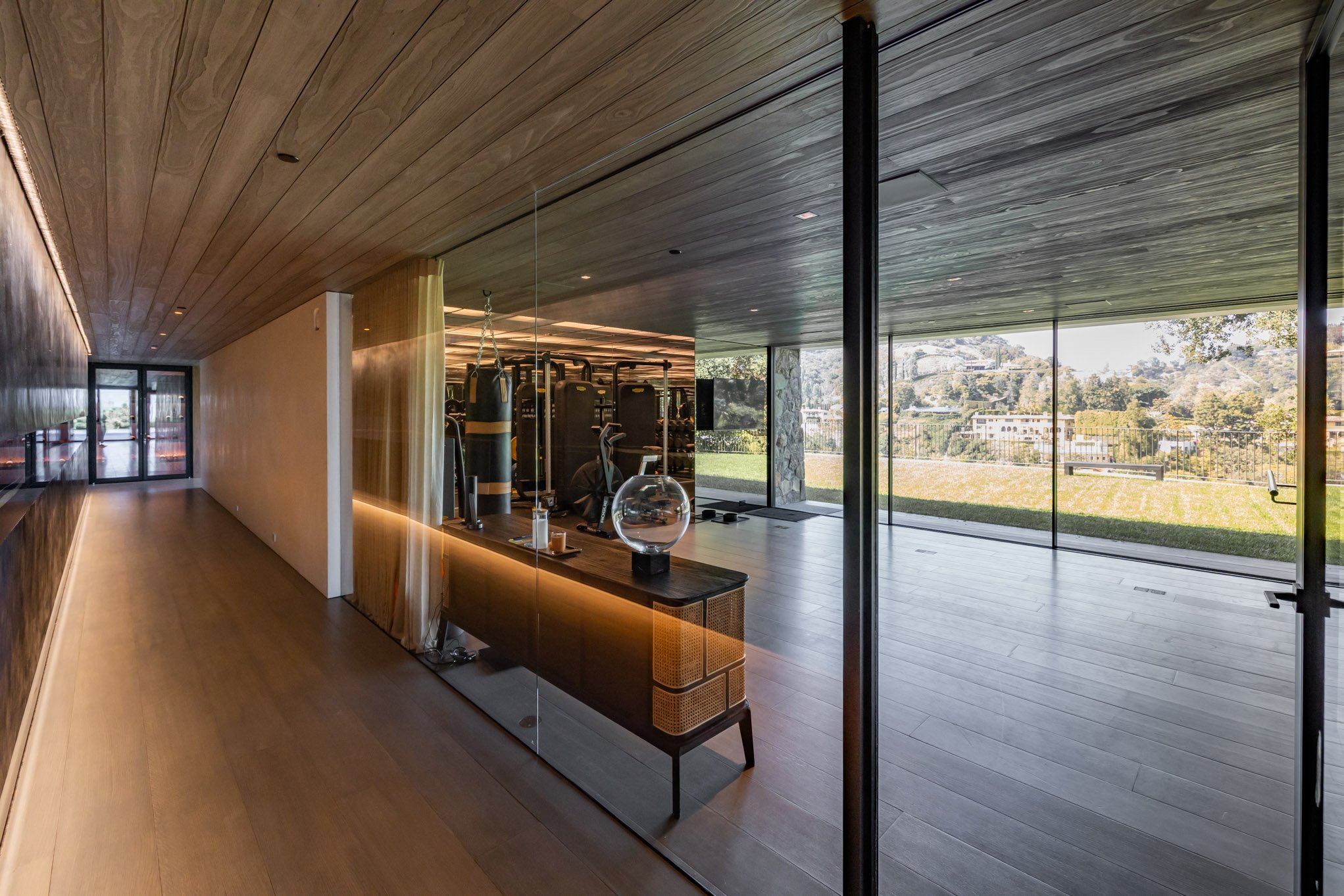Modern gym with large glass walls overlooking a grassy outdoor area, featuring elliptical trainers, punching bags, and fitness equipment inside.