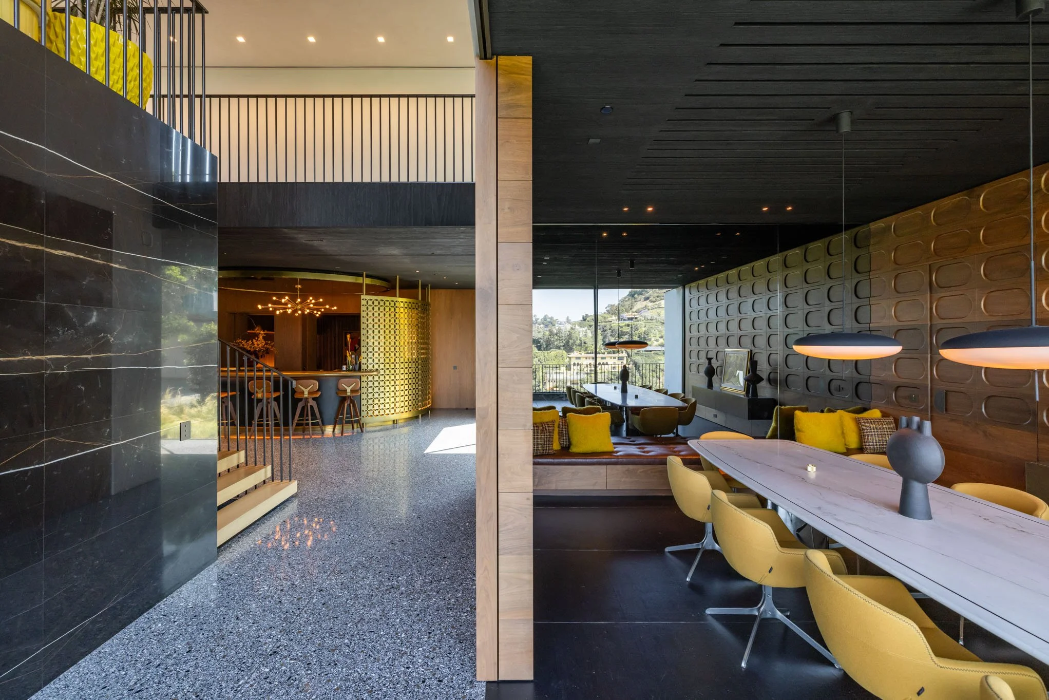 Interior of a modern, stylish home with a black marble wall, terrazzo flooring, a bright yellow bar counter, and a separate seating area with a long white table, yellow chairs, and contemporary decor.