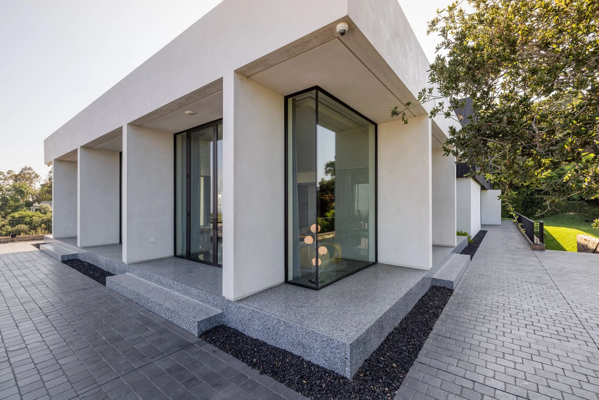 Exterior view of a modern white building with large glass windows, a paved walkway, and landscaped area with trees.