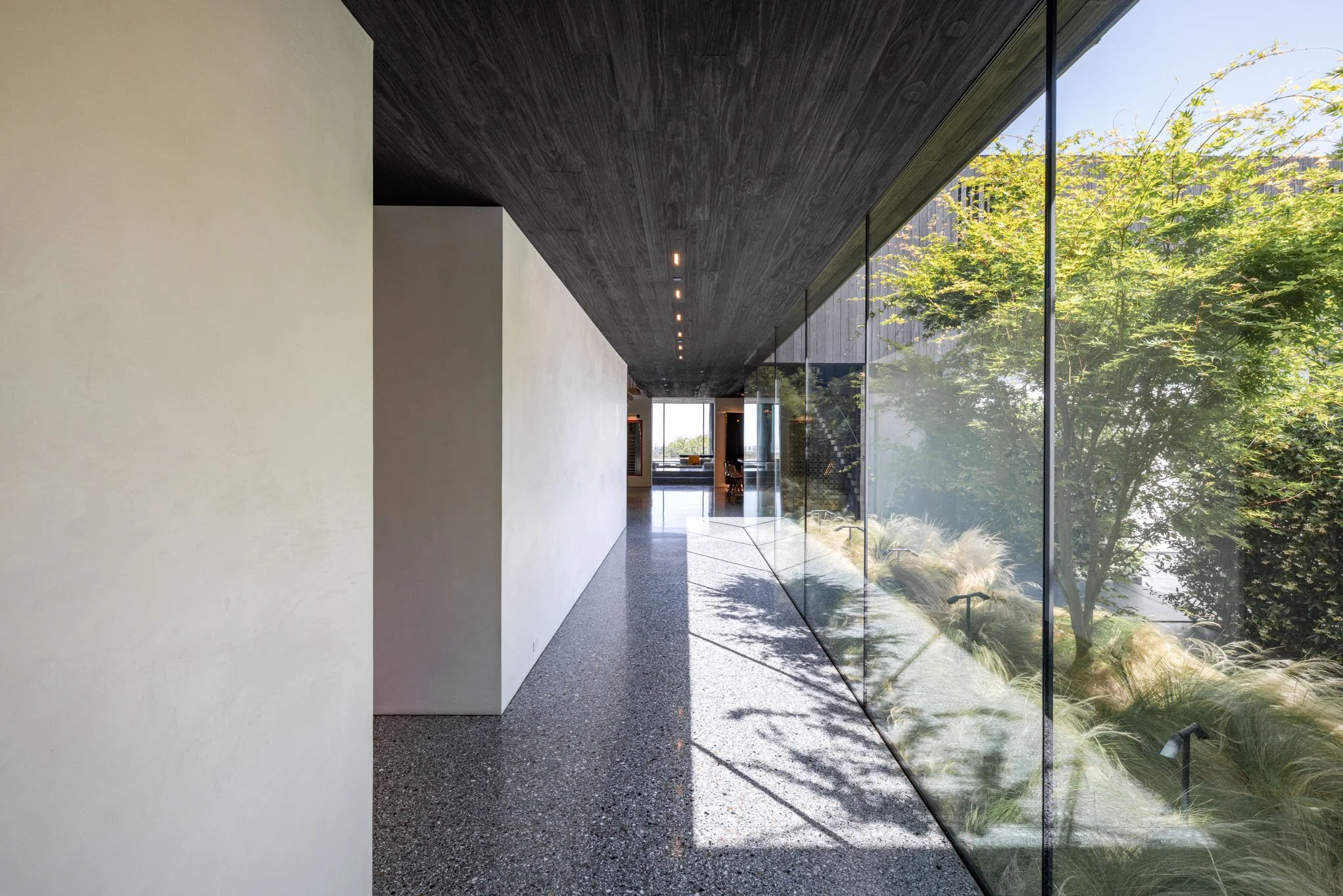 Modern hallway with stone floor, white wall on the left, dark ceiling, and large glass wall on the right showing outdoor greenery and trees.