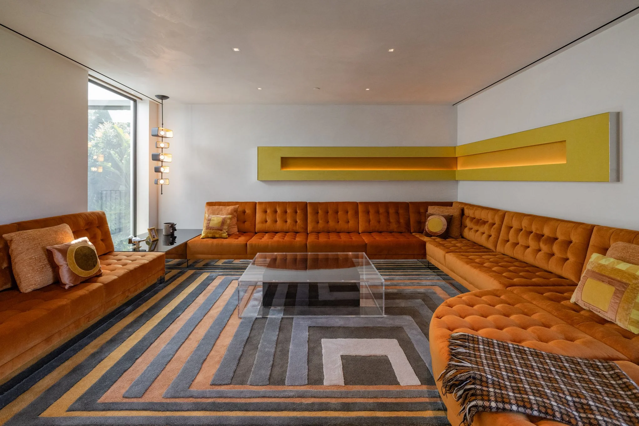 Living room with large orange sectional sofa, glass coffee table, geometric patterned rug, and modern artwork on white walls, illuminated by natural light from a large window.