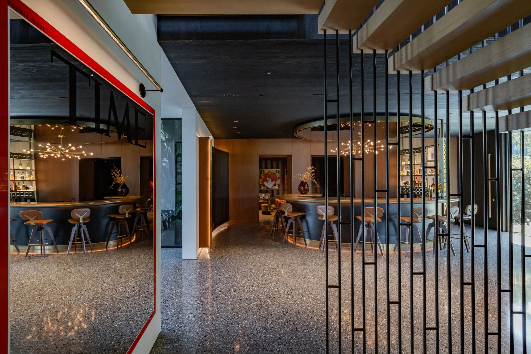 Modern bar area with a curved counter, high stools, decorative lighting fixtures, and a mix of dark wood and metallic finishes, separated by a black metal partition.