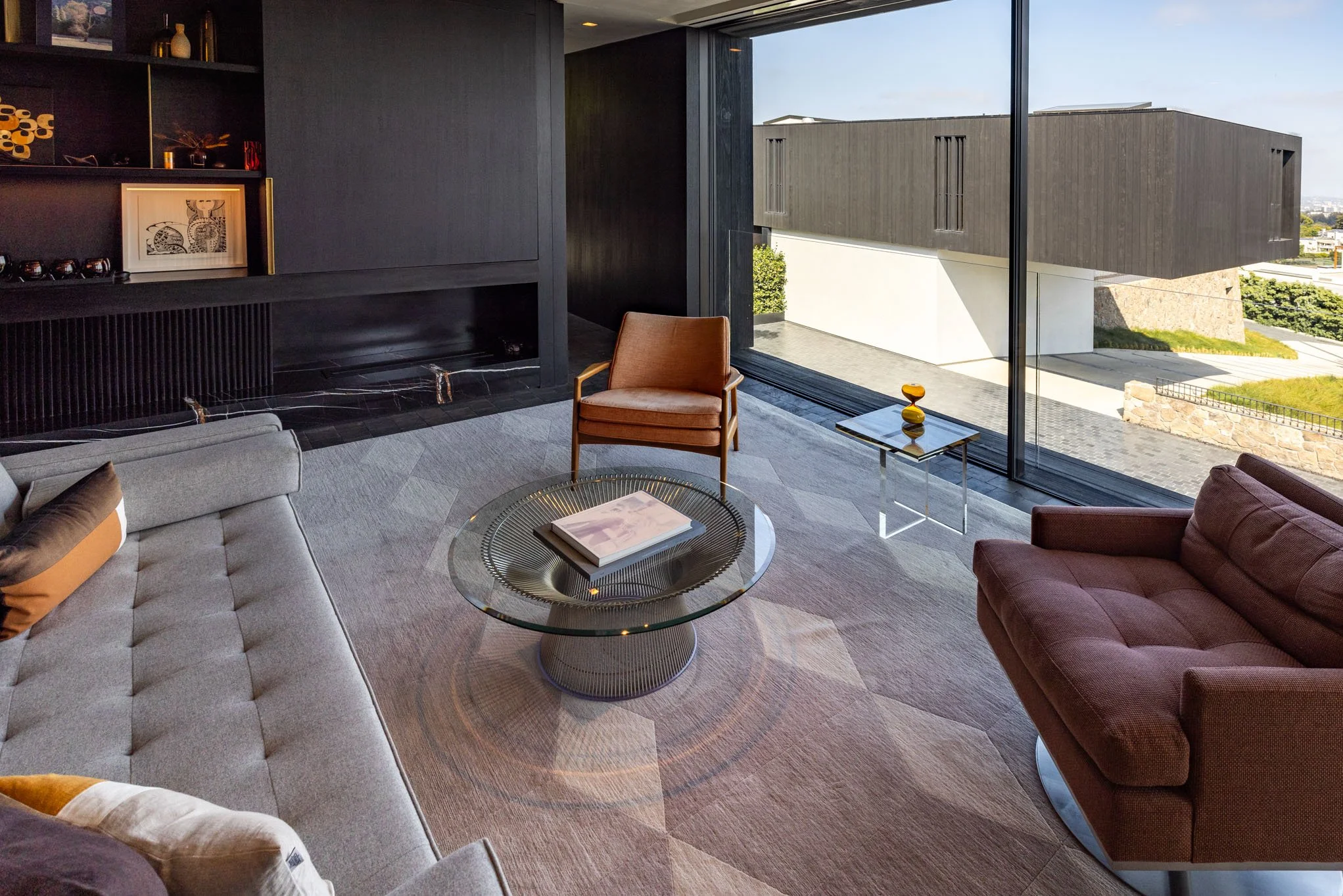 Modern living room with a large window showing an outdoor view, featuring a gray sofa, brown armchair, glass coffee table with a book, and a small side table with a decorative object. Black wall paneling and built-in shelving are visible.