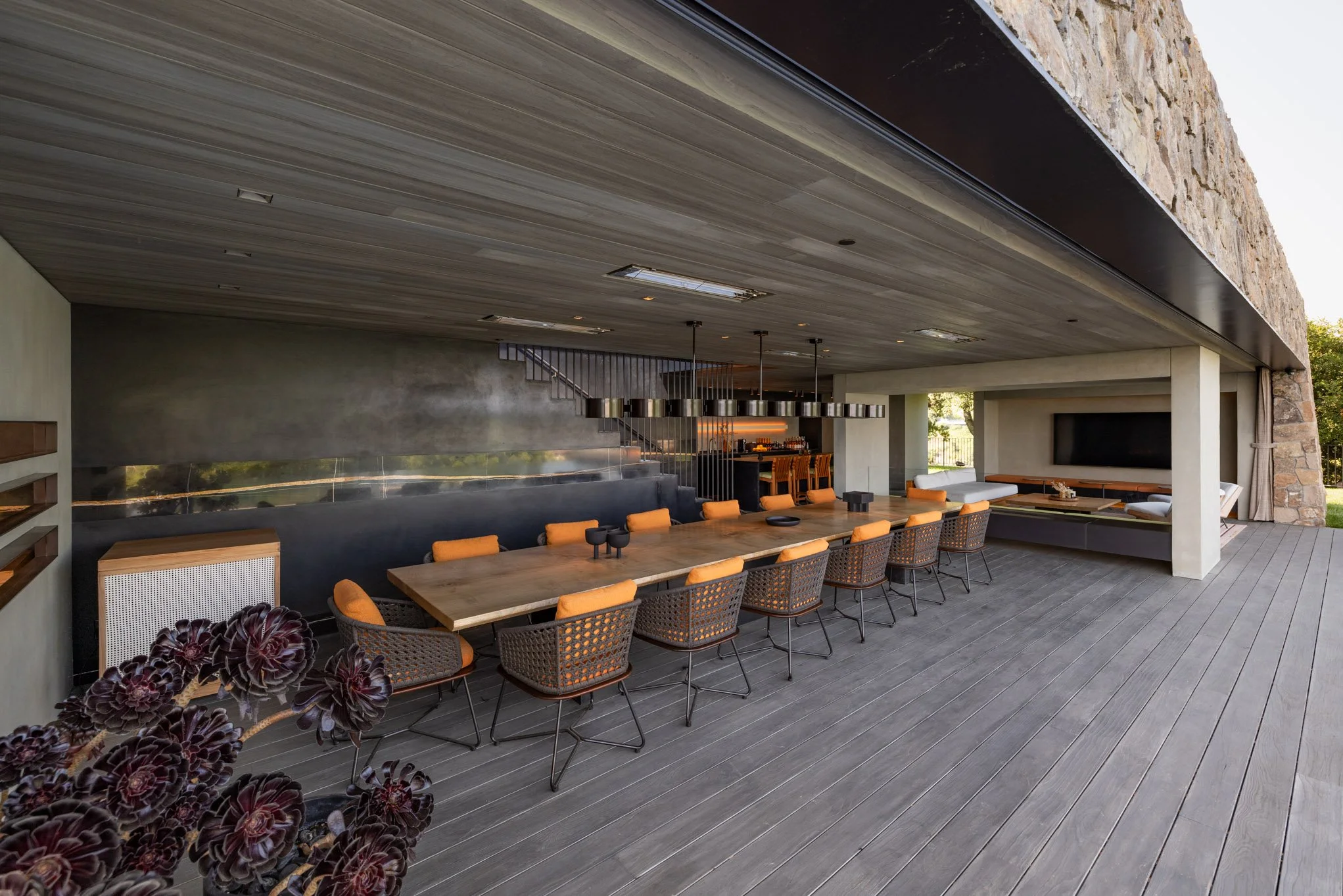 Modern outdoor patio with a large dining table, orange cushions on chairs, a built-in lounge area, and a bar in the background.