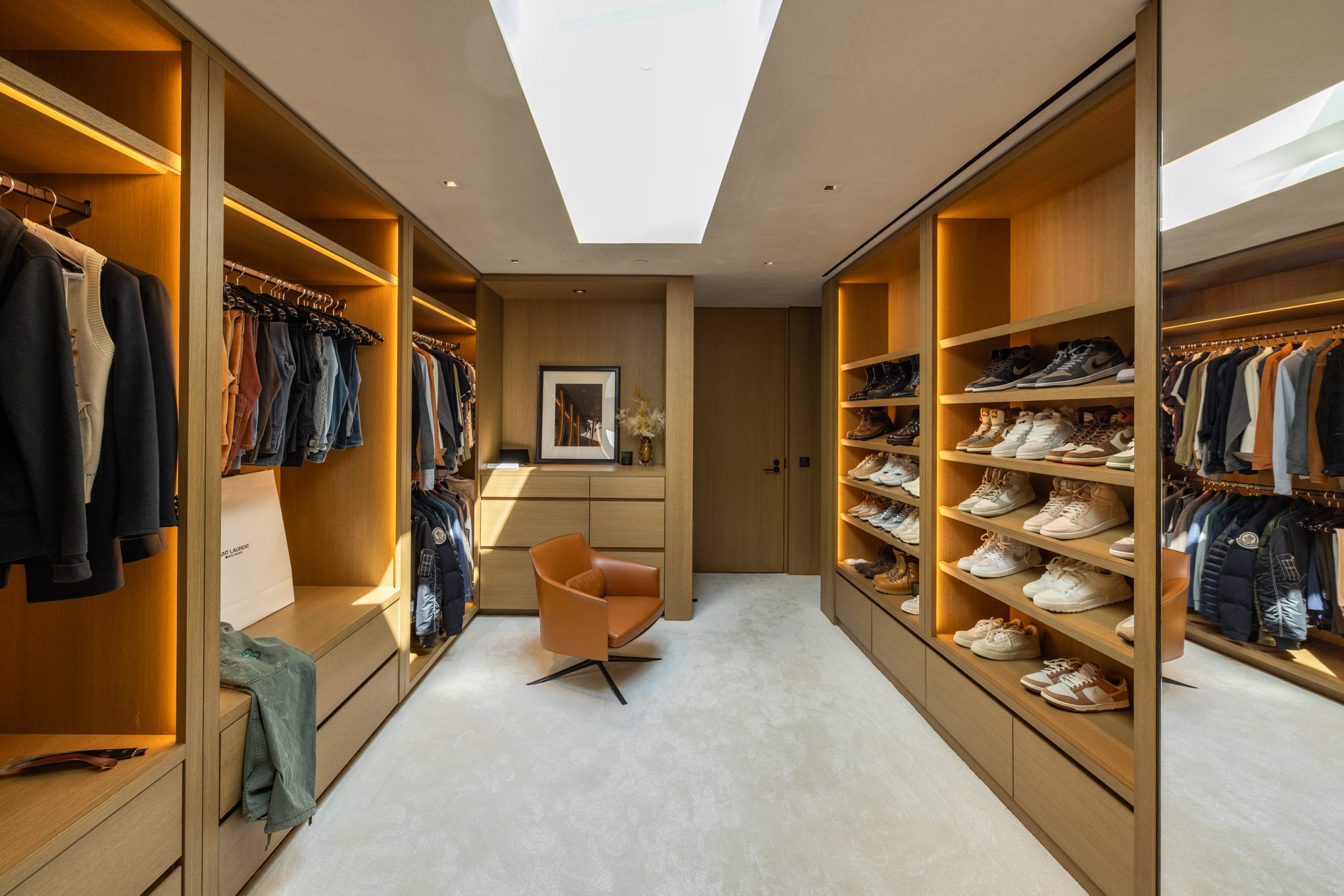 Luxurious walk-in closet with wooden shelving, clothing, shoes, a leather armchair, a framed picture, and a skylight ceiling.