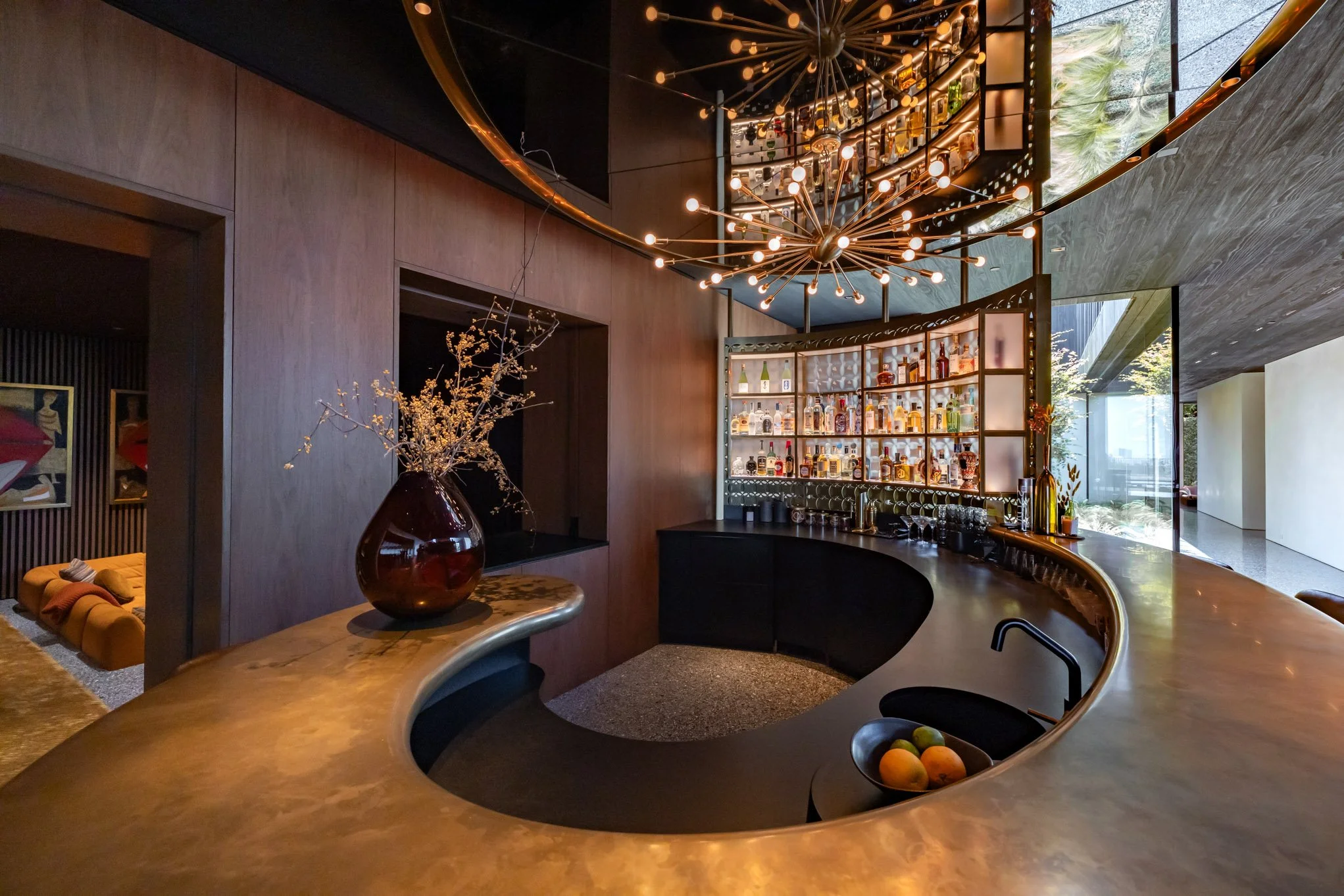Elegant modern bar with curved gold countertop, a large decorative chandelier, and a backlit bottle display with barware and glassware. A vase with dried flowers is on the counter, and a bowl of fruit is near the sink. Large windows reveal greenery o