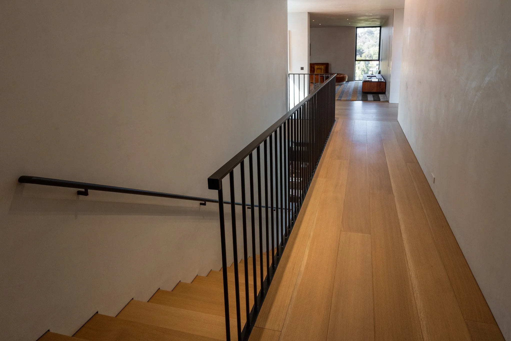 Interior view of a modern house hallway with wooden flooring, a staircase with a black metal railing, and a living room with a large window and minimal furniture.